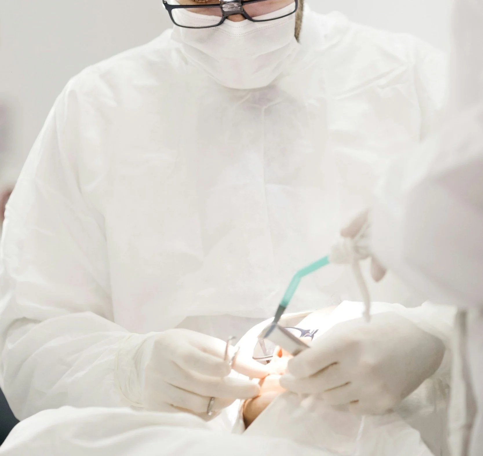 A medical professional in white protective clothing, gloves, a face mask, and glasses performing a procedure with medical instruments.
