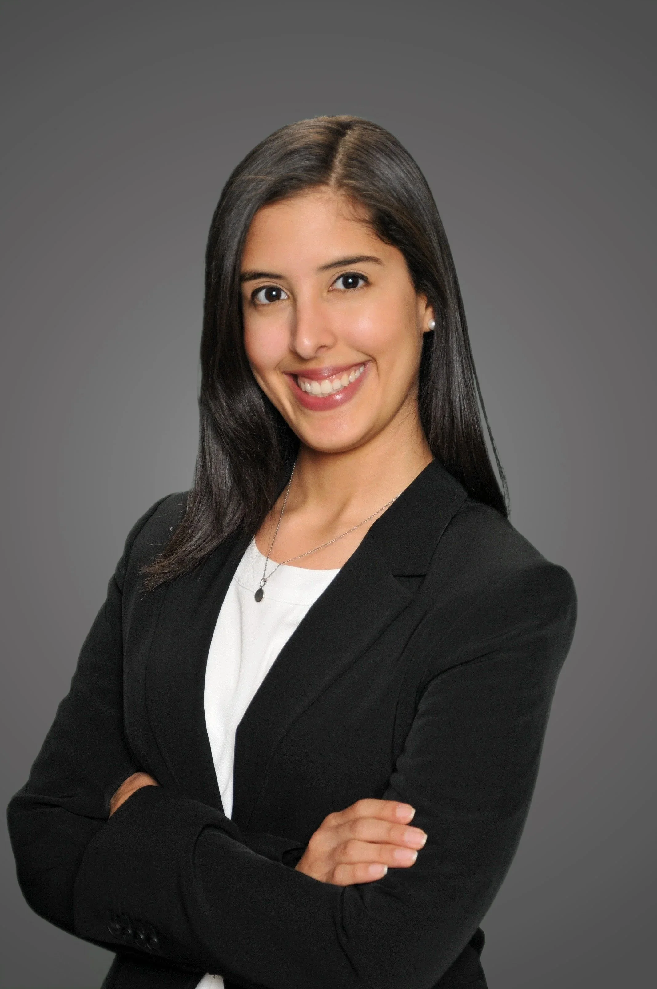 Professional woman with long dark hair, wearing a black blazer and white top, smiling with arms crossed against a gray background.