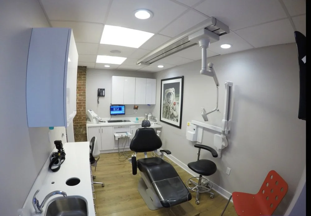 A medical examination room with a dental chair, medical equipment, white cabinets, a computer, and medical tools. There's a framed picture on the wall and ceiling lights.