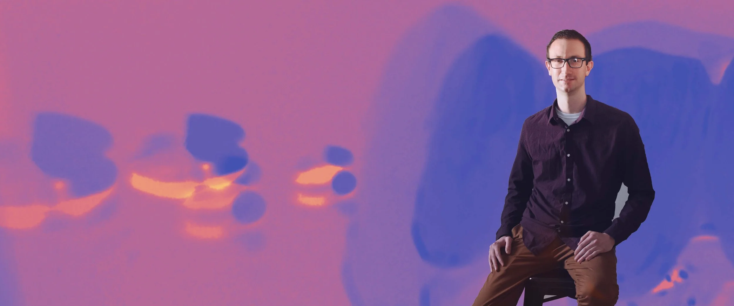 A man with glasses and a dark shirt sitting on a stool with a colorful abstract background.