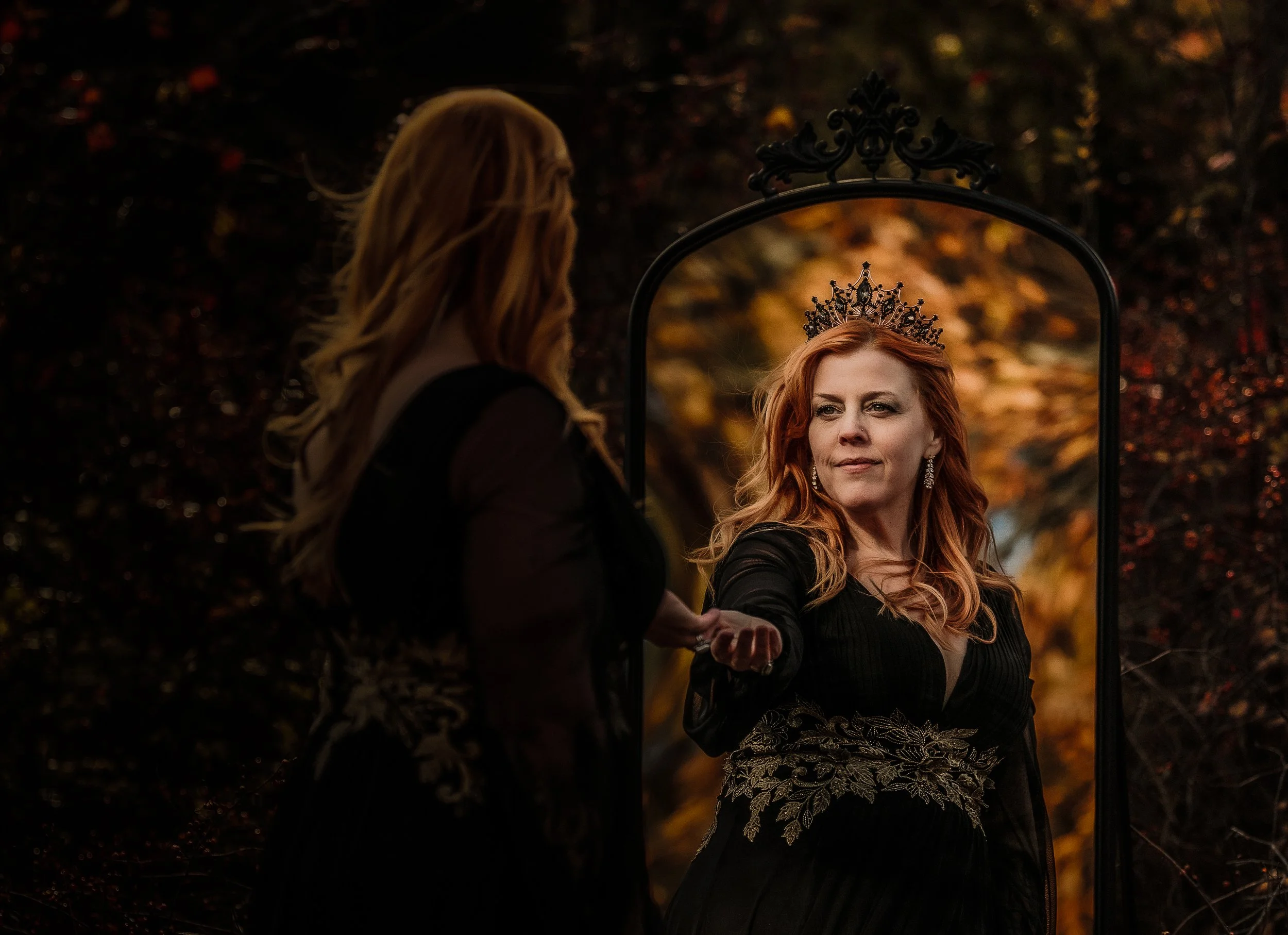 A woman with red hair wearing a crown looks at herself in a mirror outdoors during autumn. She wears a black dress with gold embroidery and earrings, and holds a hand out toward the mirror.