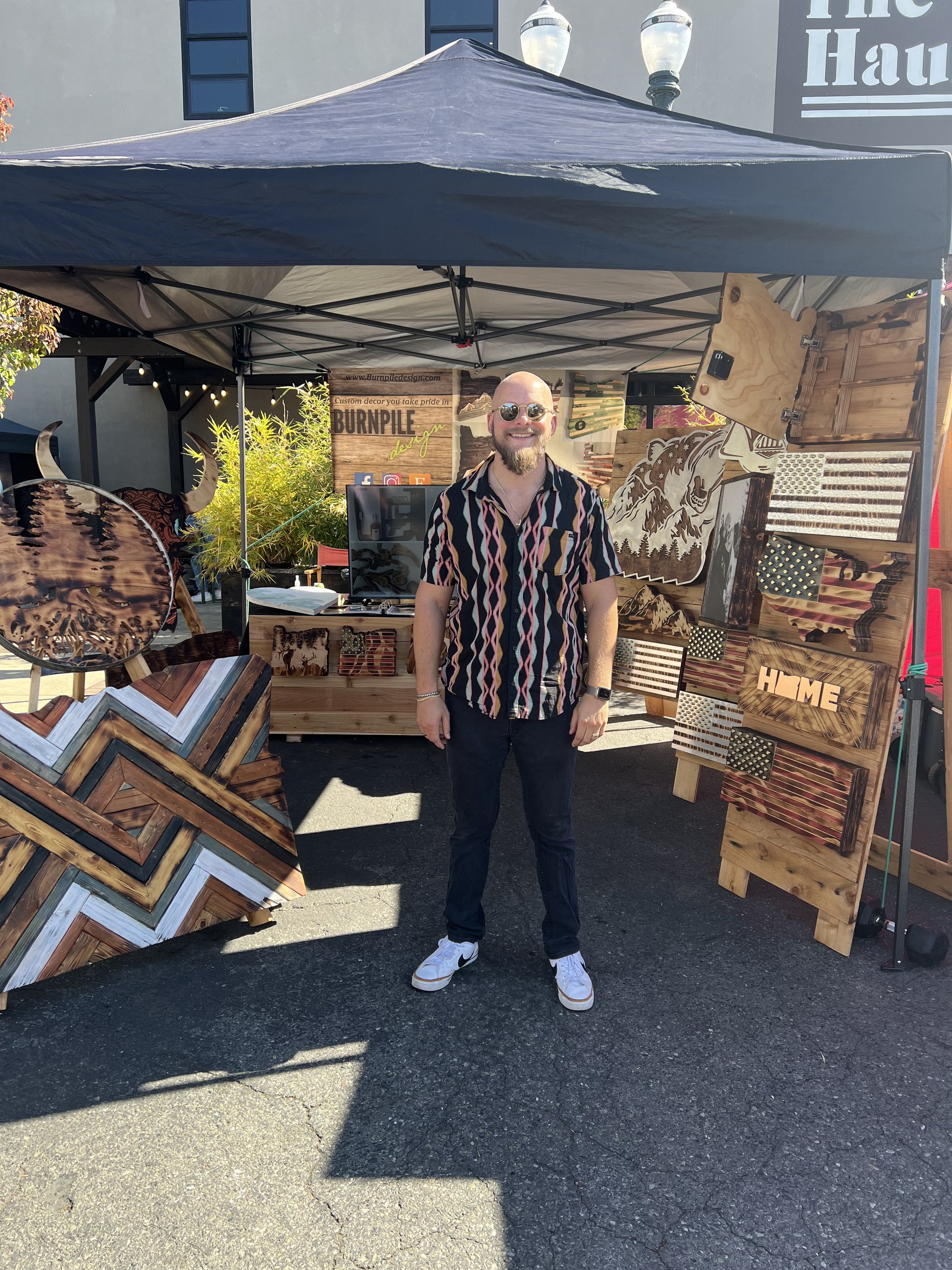 A man standing in front of a booth at an outdoor market. The booth is covered by a black canopy and displays various wooden art pieces and decorations, including a geometric design and wood burn art, one with the word "HOME." The man is wearing sunglasses, a striped shirt, jeans, and white sneakers. The setting suggests a craft fair or market.