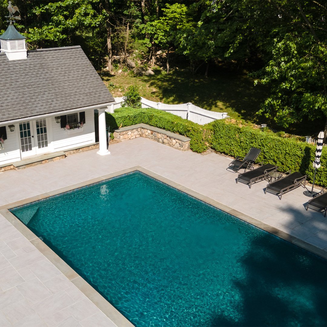 An elevated view of a stunning outdoor retreat.
This expansive Light Gray Porcelain patio is designed for comfort, relaxation, and enjoying the outdoors. Featuring a Bluestone coping pool and a Bluestone-capped Fieldstone wall, this private backyard 