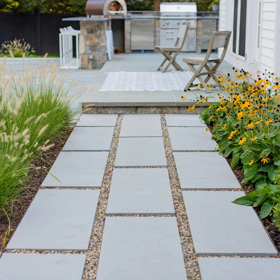 A seamless outdoor escape! Bluestone flagging with New Jersey Beach Gravel leads the way to a beautifully designed Bluestone patio, complete with chairs for relaxing and an outdoor kitchen perfect for entertaining.