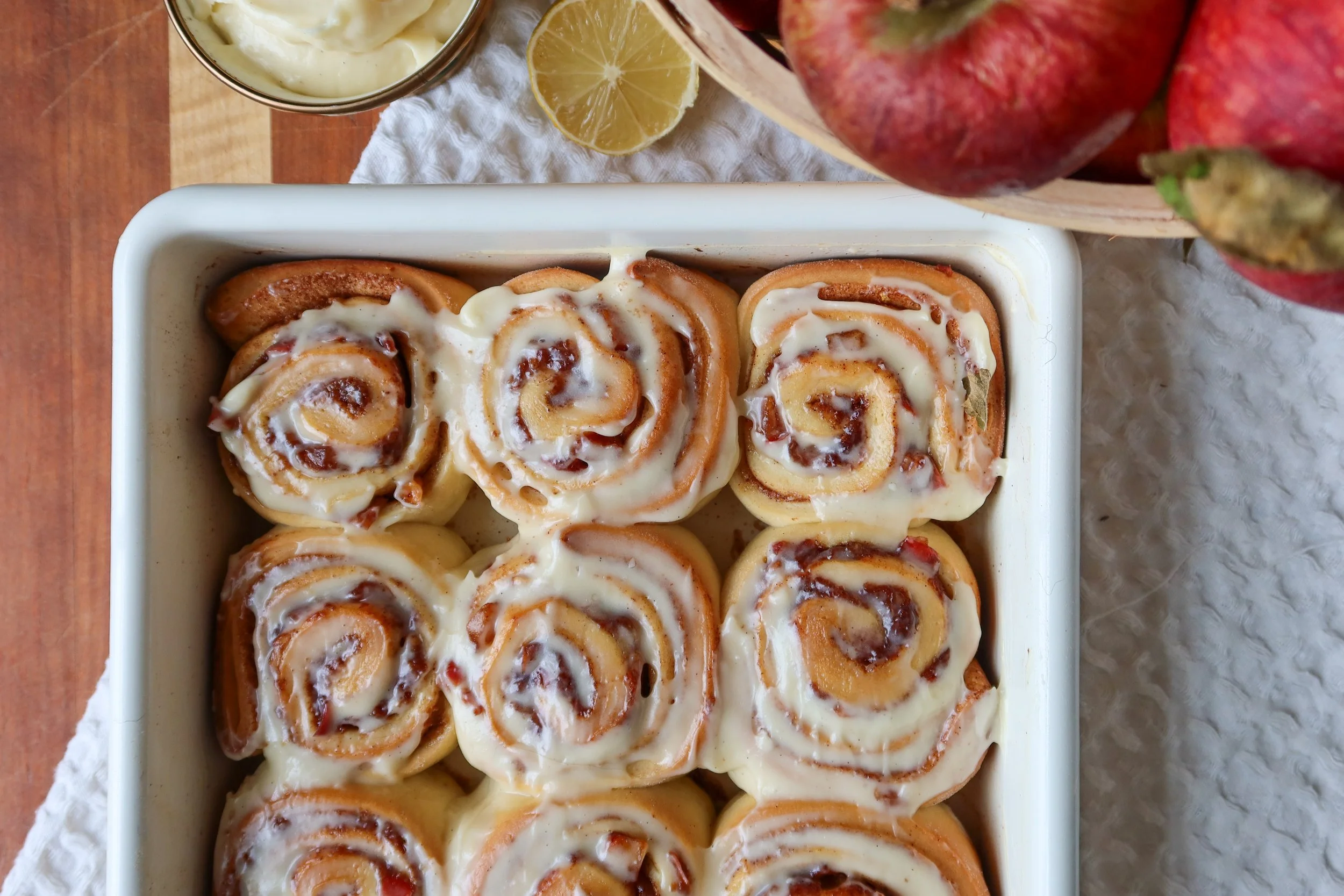 Apple-filled cinnamon rolls with maple cream cheese glaze in a MacKenzie-Childs baking dish.