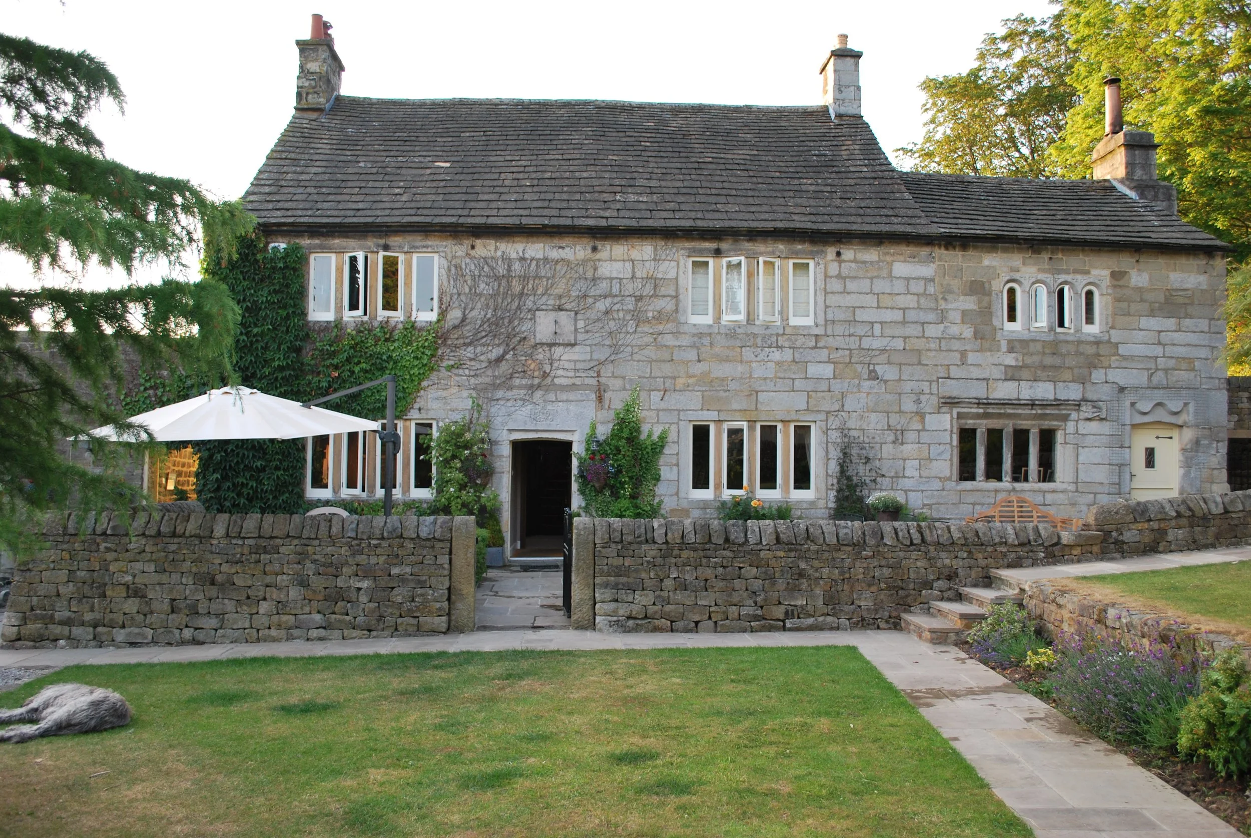 A stone house with multiple white-framed windows, surrounded by a low stone wall, with a garden and a pathway leading to the front door, and an outdoor umbrella on the left side.