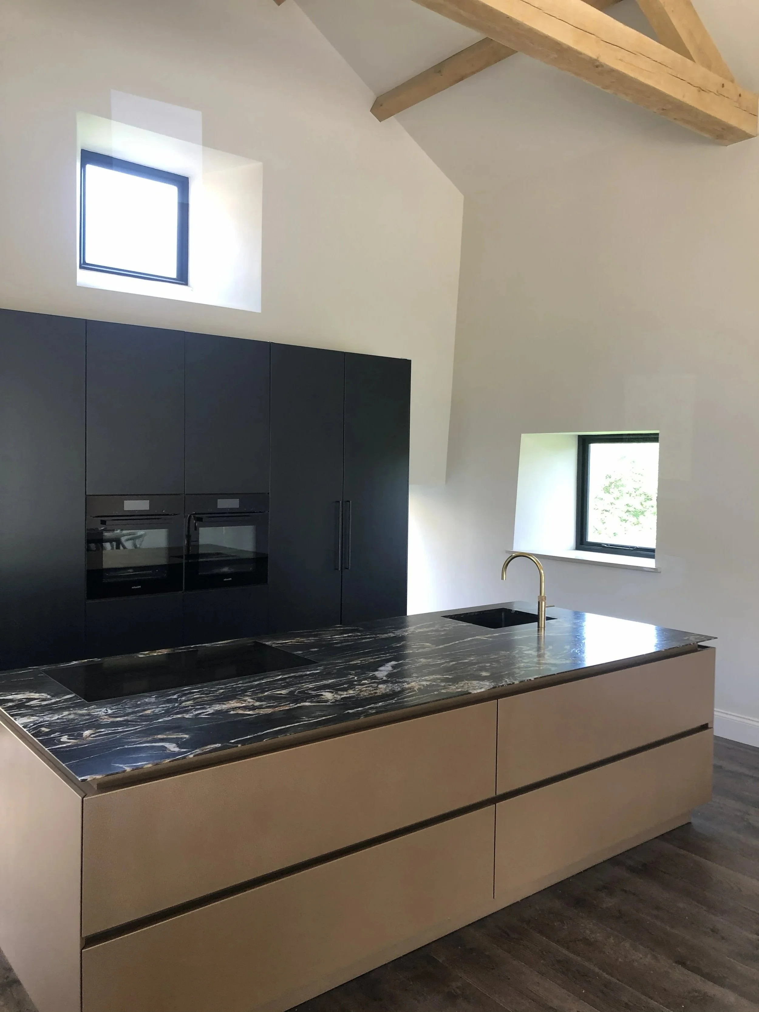 Modern kitchen with black cabinetry, marble countertop island, gold faucet, and two small windows in a bright, minimalist space with wooden beams.