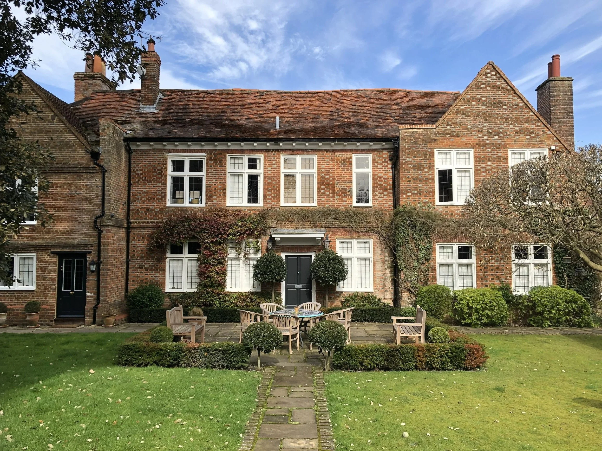 A large brick house with multiple windows, a black front door, surrounded by a garden with trimmed bushes, a stone pathway, and outdoor seating under a blue sky.