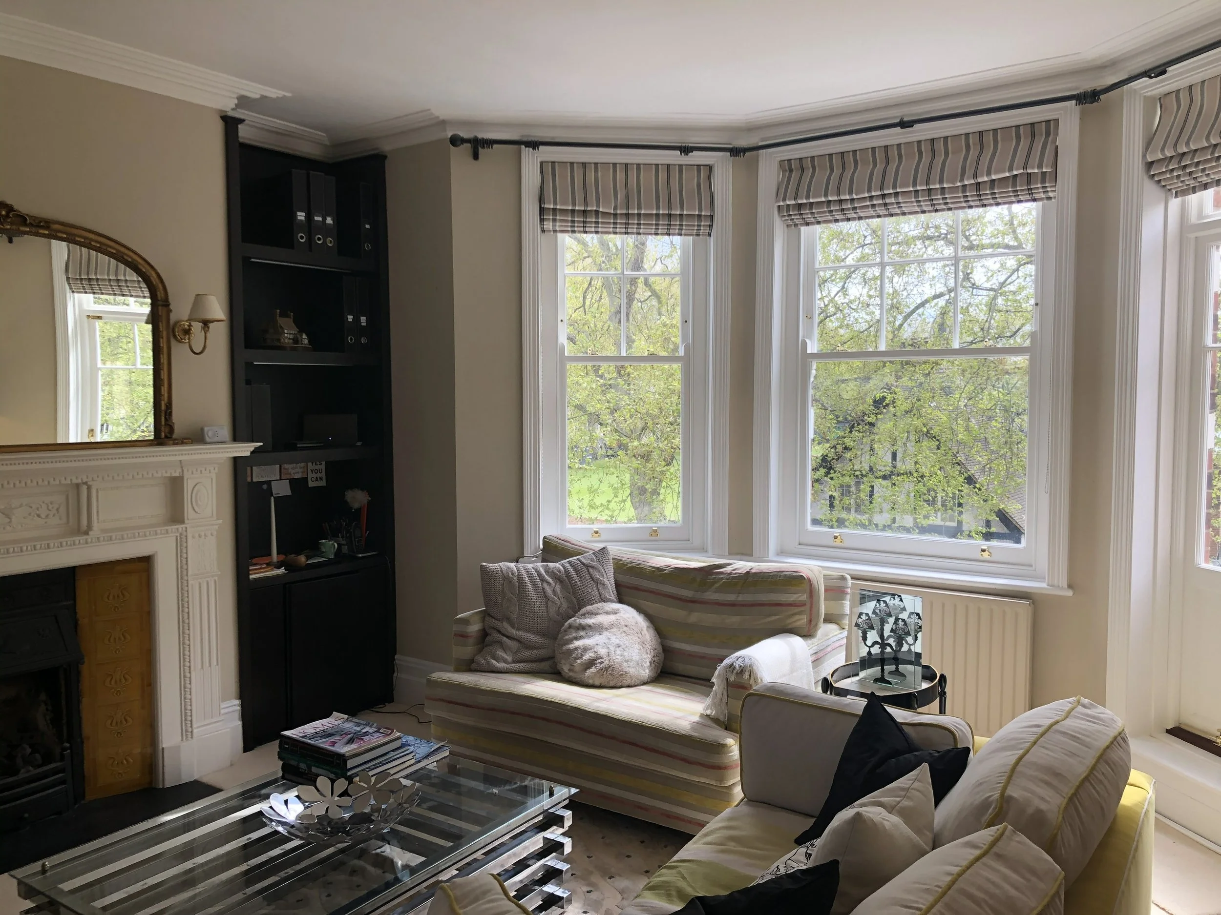 Bright living room with large bay windows, a yellow couch with throw pillows, a striped armchair, a glass coffee table with a metal decorative bowl, and a black bookshelf next to a fireplace with a mirror above.