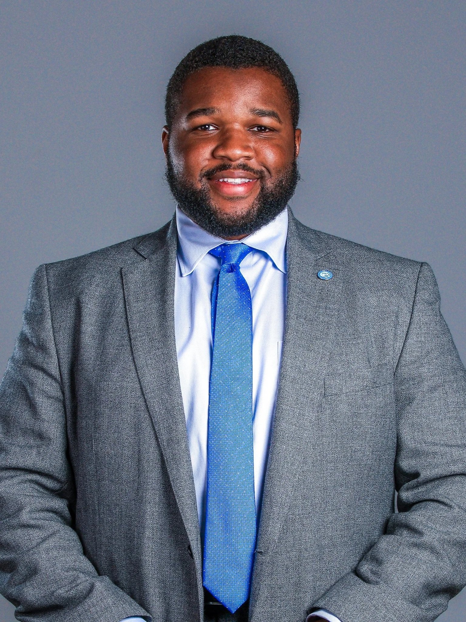 A cheerful middle-aged African American man wearing a light pink plaid blazer over a red shirt, with arms crossed, smiling against a gray background.