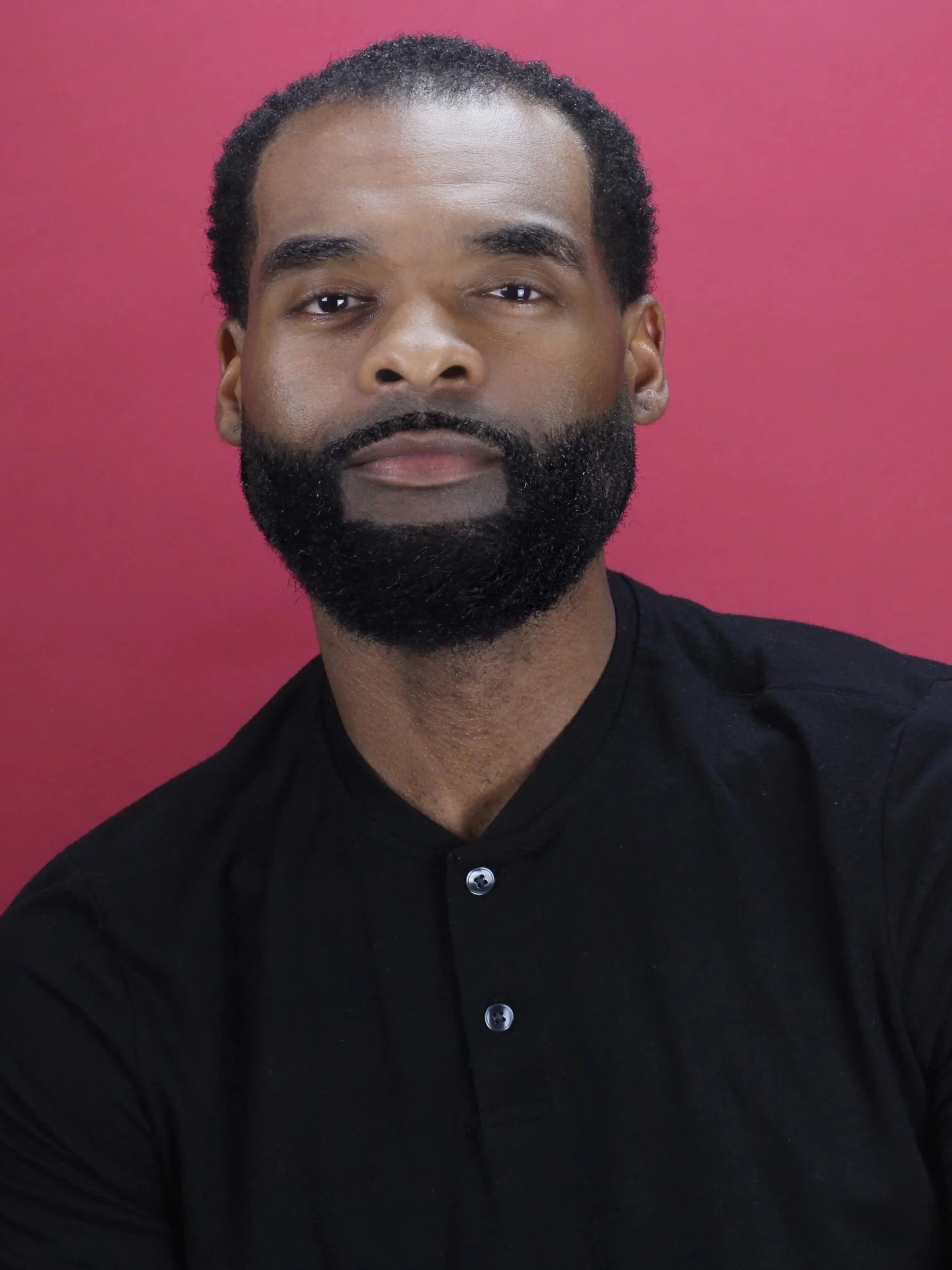 A cheerful middle-aged African American man wearing a light pink plaid blazer over a red shirt, with arms crossed, smiling against a gray background.