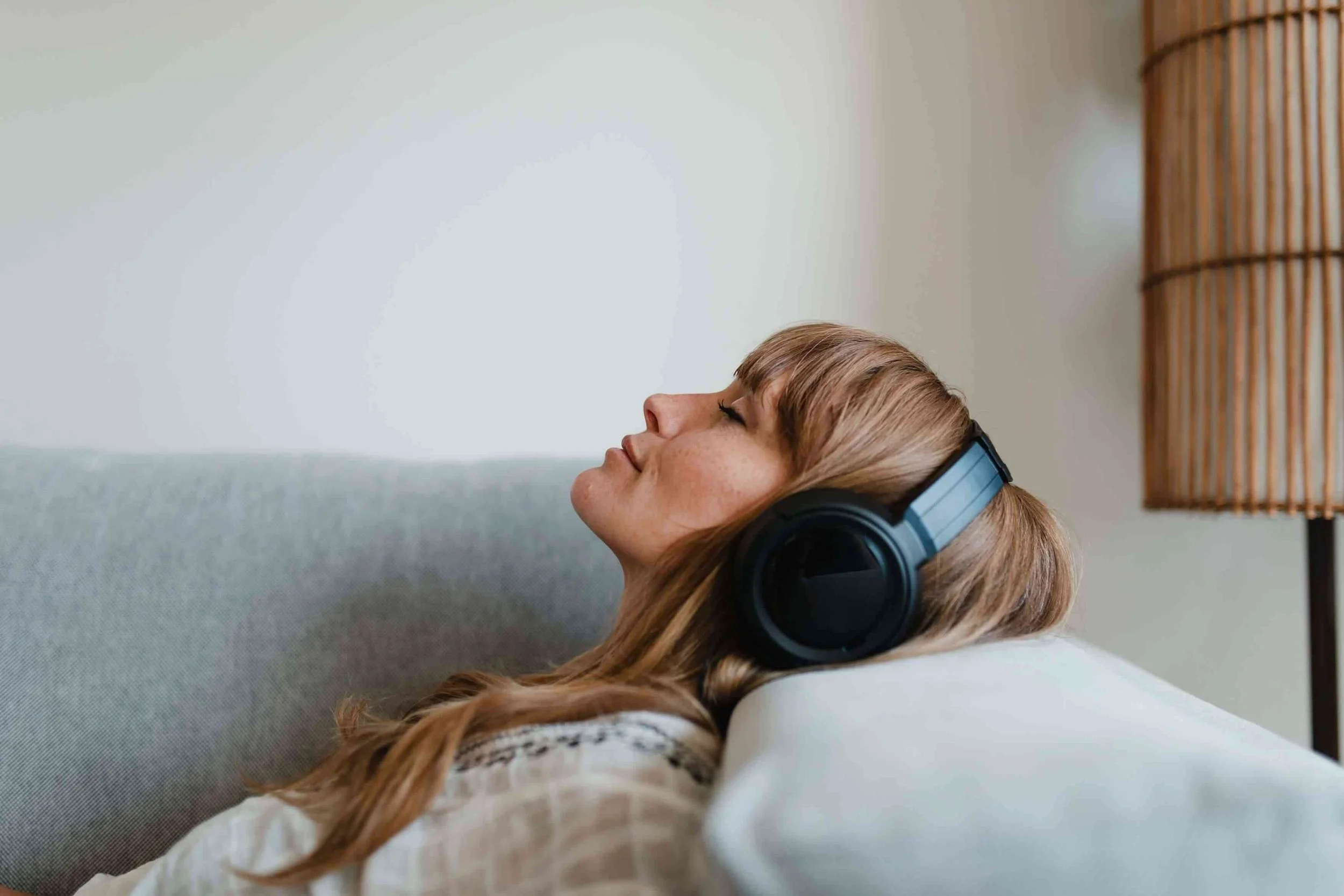 Woman with long hair wearing headphones and resting her head on a cushion with her eyes closed in a cozy room.