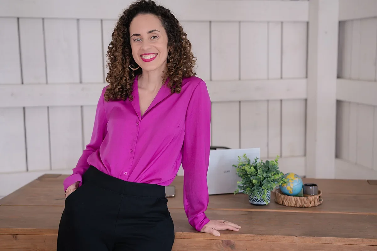 Katrina McGhee in magenta blouse and black pants leaning on desk in bright studio for sabbatical coaching