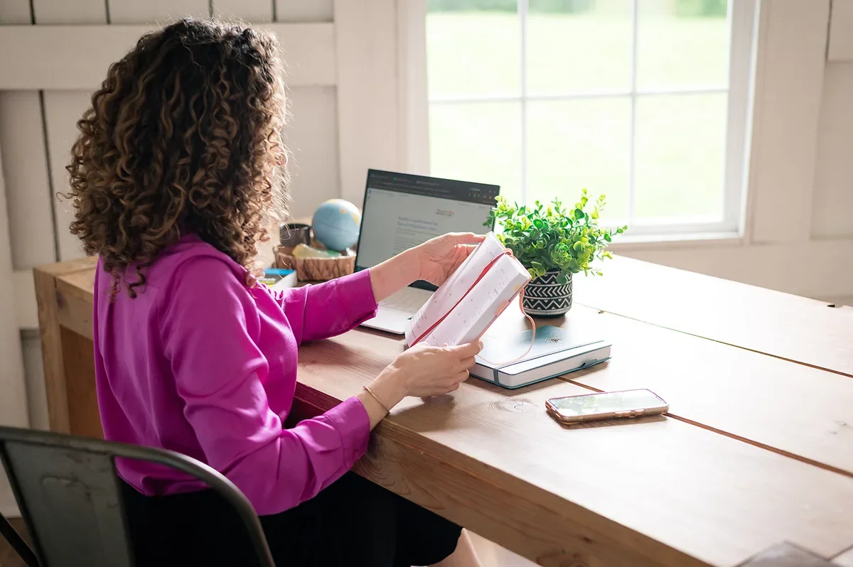 Woman in pink blouse reading journal at wooden desk with laptop, globe, and window light