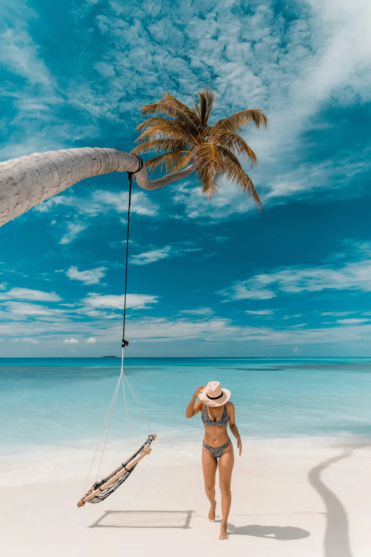 Woman in bikini walking on a white sand beach beneath a leaning palm tree with a rope swing, evoking career break adventure