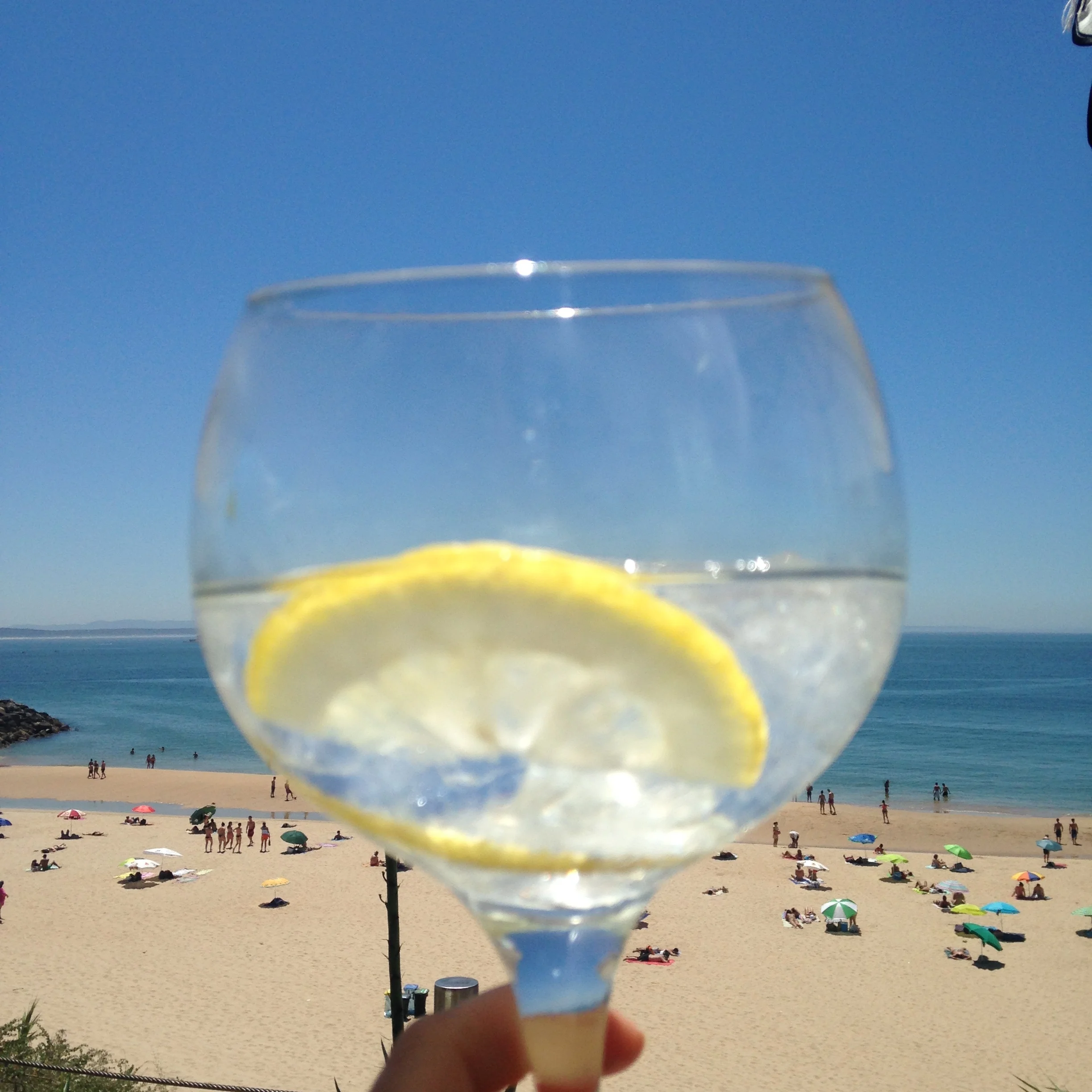 A slice of lemon in a glass held up, with a beach and ocean in the background. The beach has people, umbrellas, and a clear blue sky.