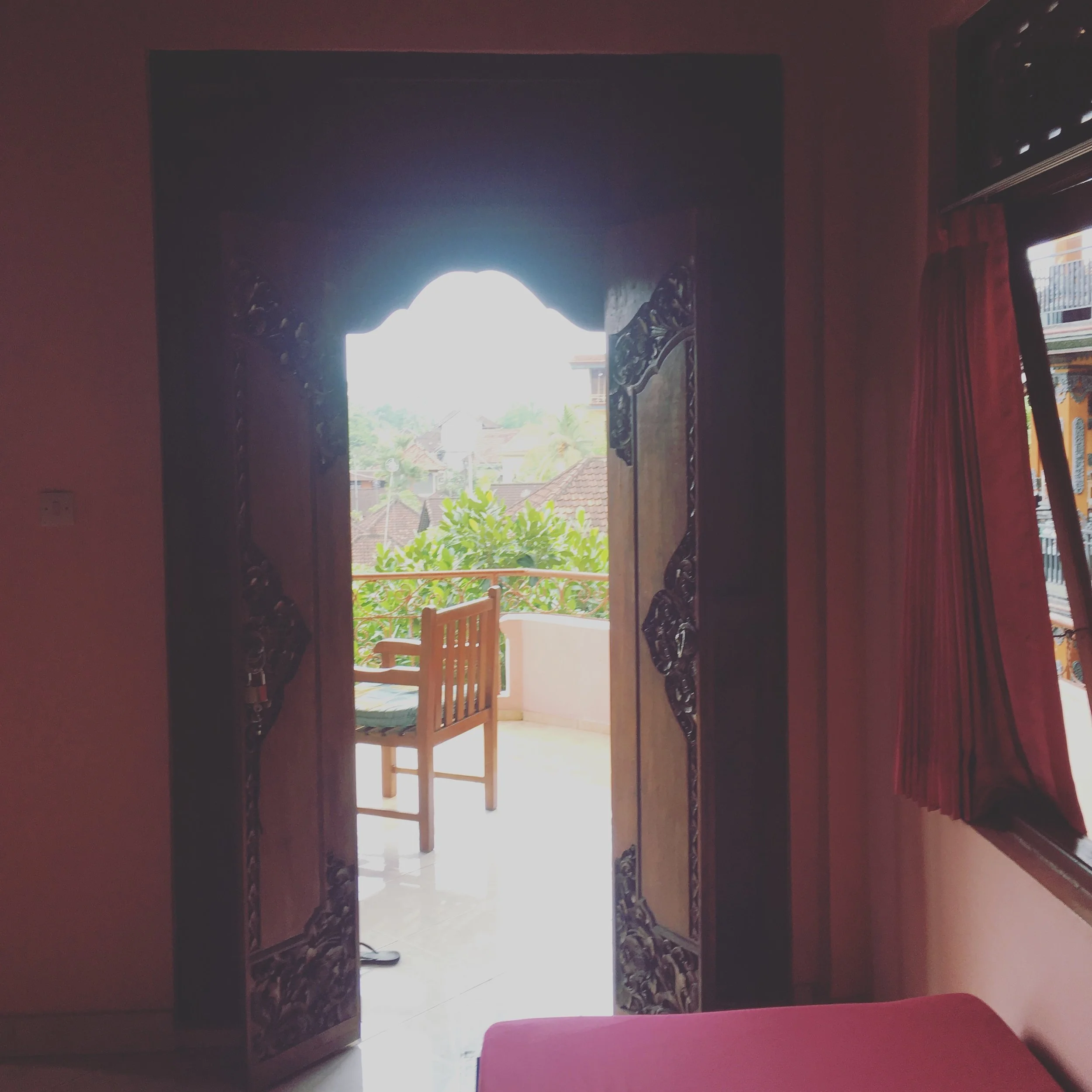 View through an ornate wooden doorway to a balcony with a wooden chair and greenery outside.
