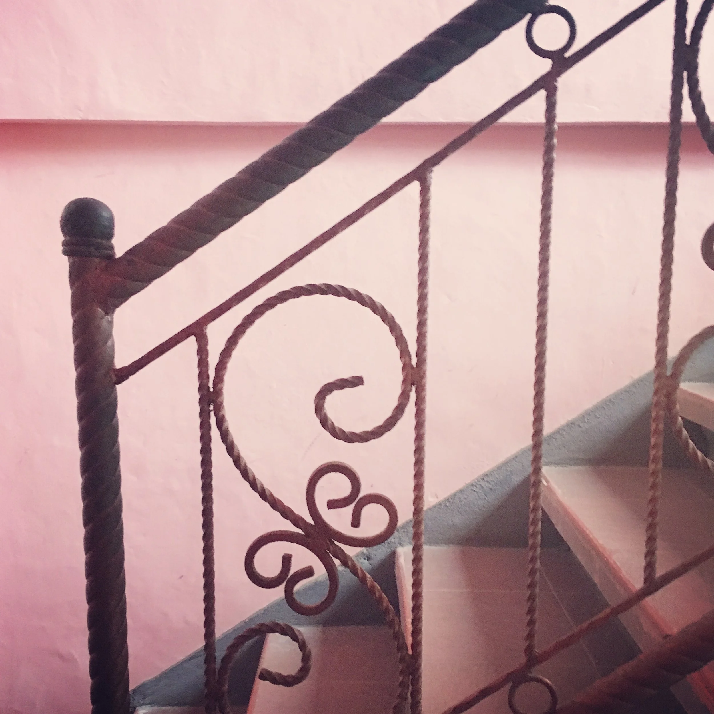 Close-up of an ornate wrought iron staircase railing with decorative scrollwork against a pink wall.