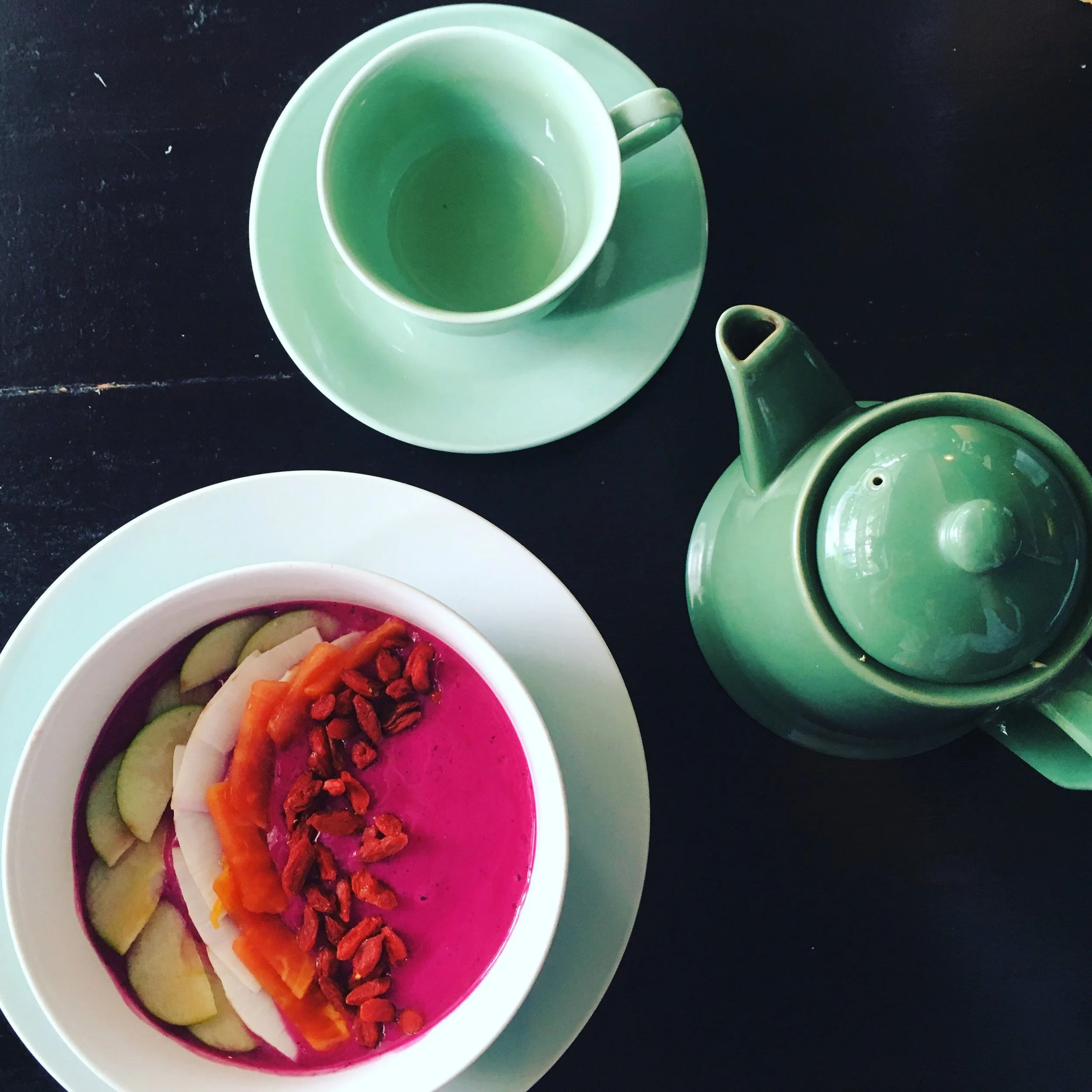 A vibrant pink smoothie bowl with sliced fruits and goji berries, accompanied by a green teacup and teapot on a dark surface.