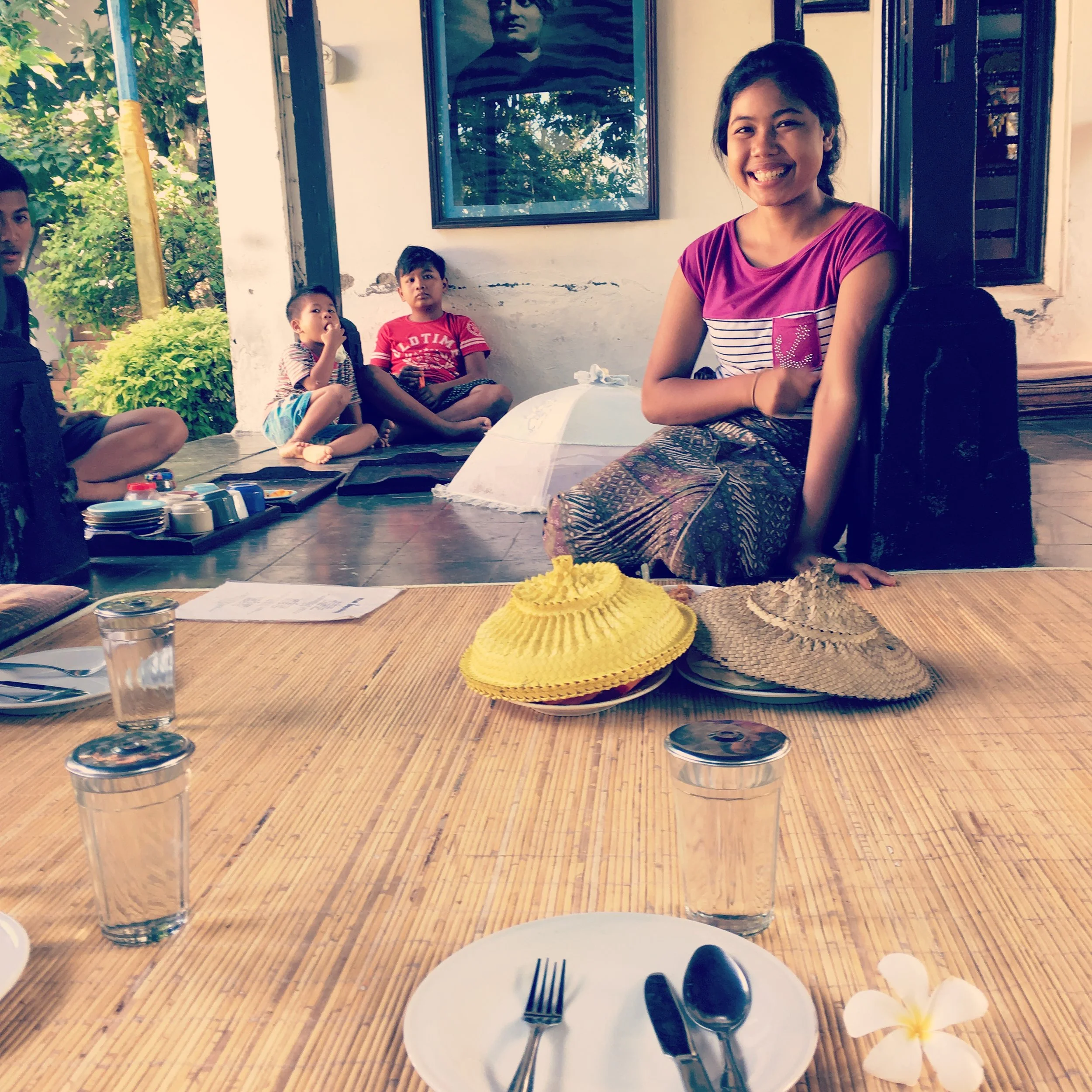 Group of people sitting indoors on wooden floor, plates and covered dishes on a mat, with cups of water, and a person smiling in the background.