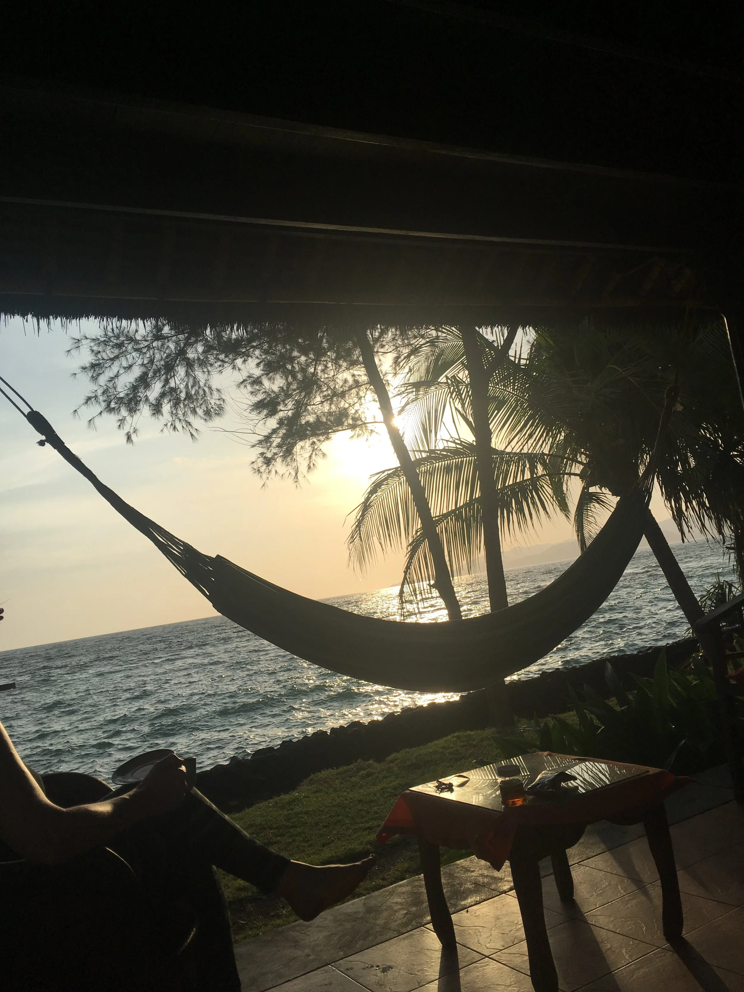 Silhouette of a person sitting near a hammock by the sea with palm trees and a sunset background.