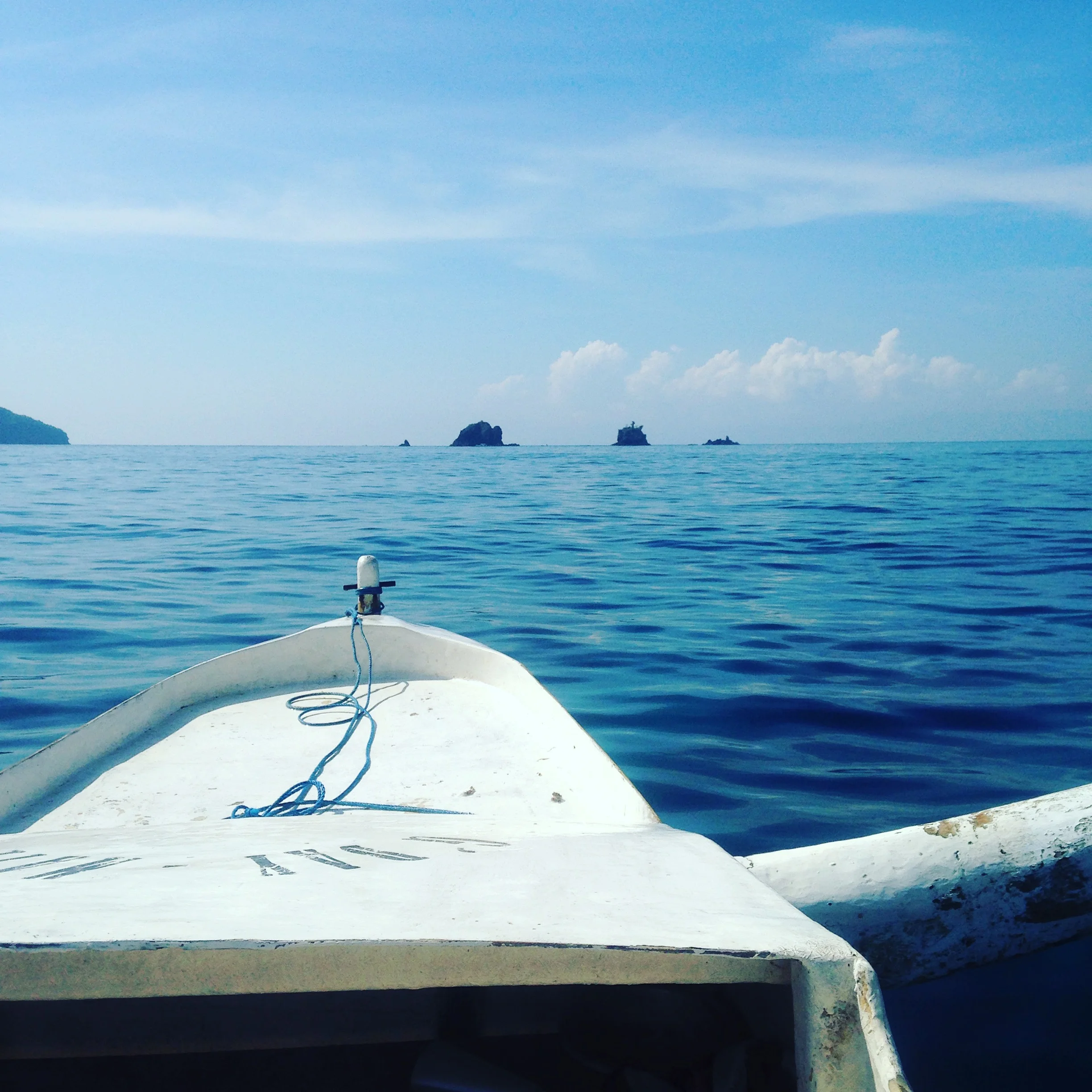 View from the bow of a small boat on a calm ocean, with distant rocky islands and a clear blue sky.