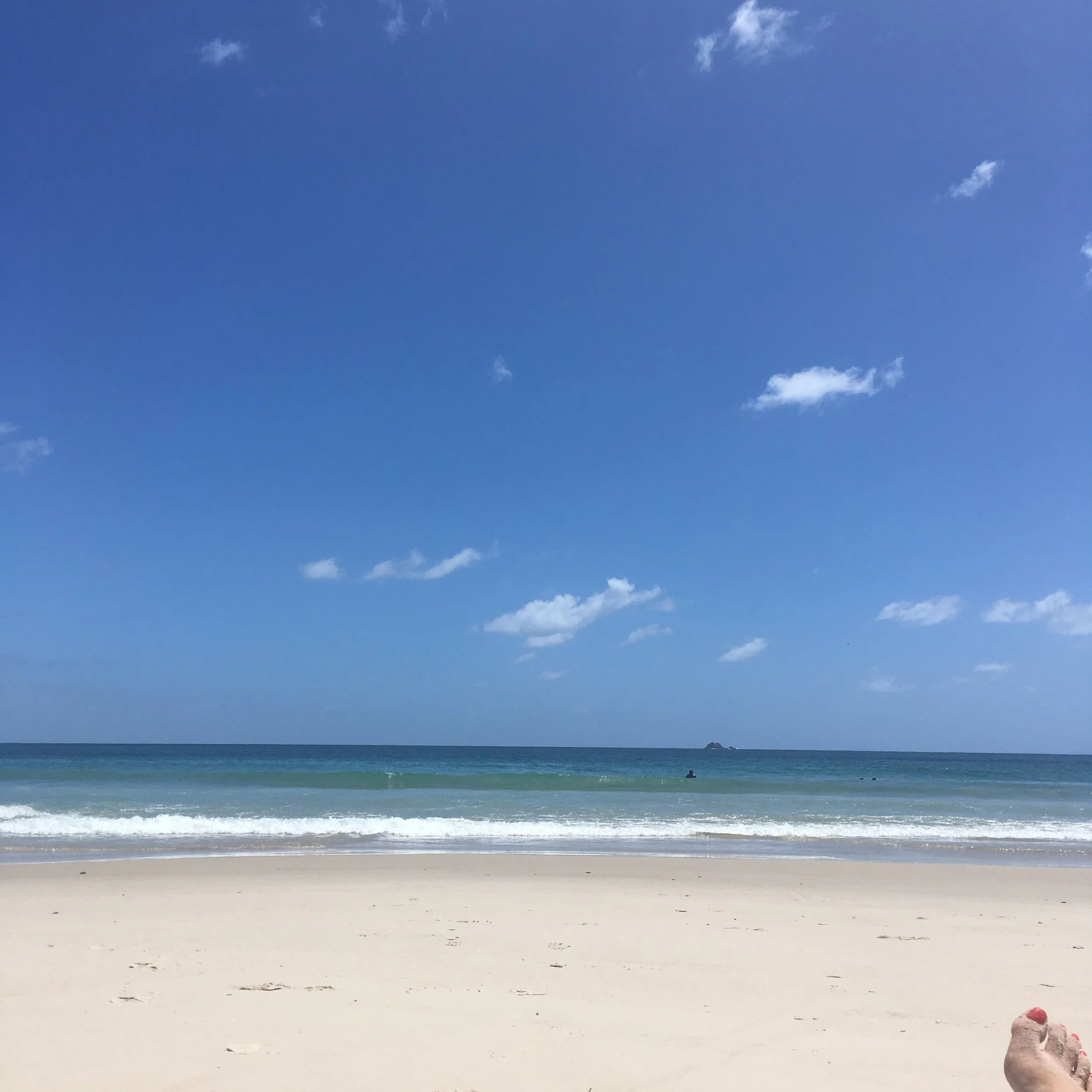 Sunny beach view with clear blue sky, ocean waves, sandy shore, and a small rock in the distance.