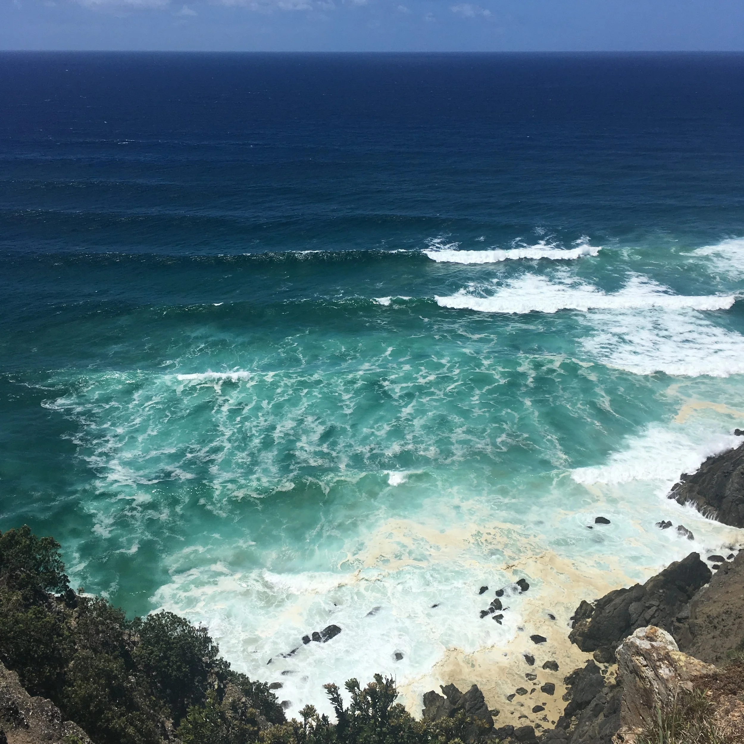 Coastal view with waves crashing on rocky shore, blue ocean extending to the horizon under a clear sky.