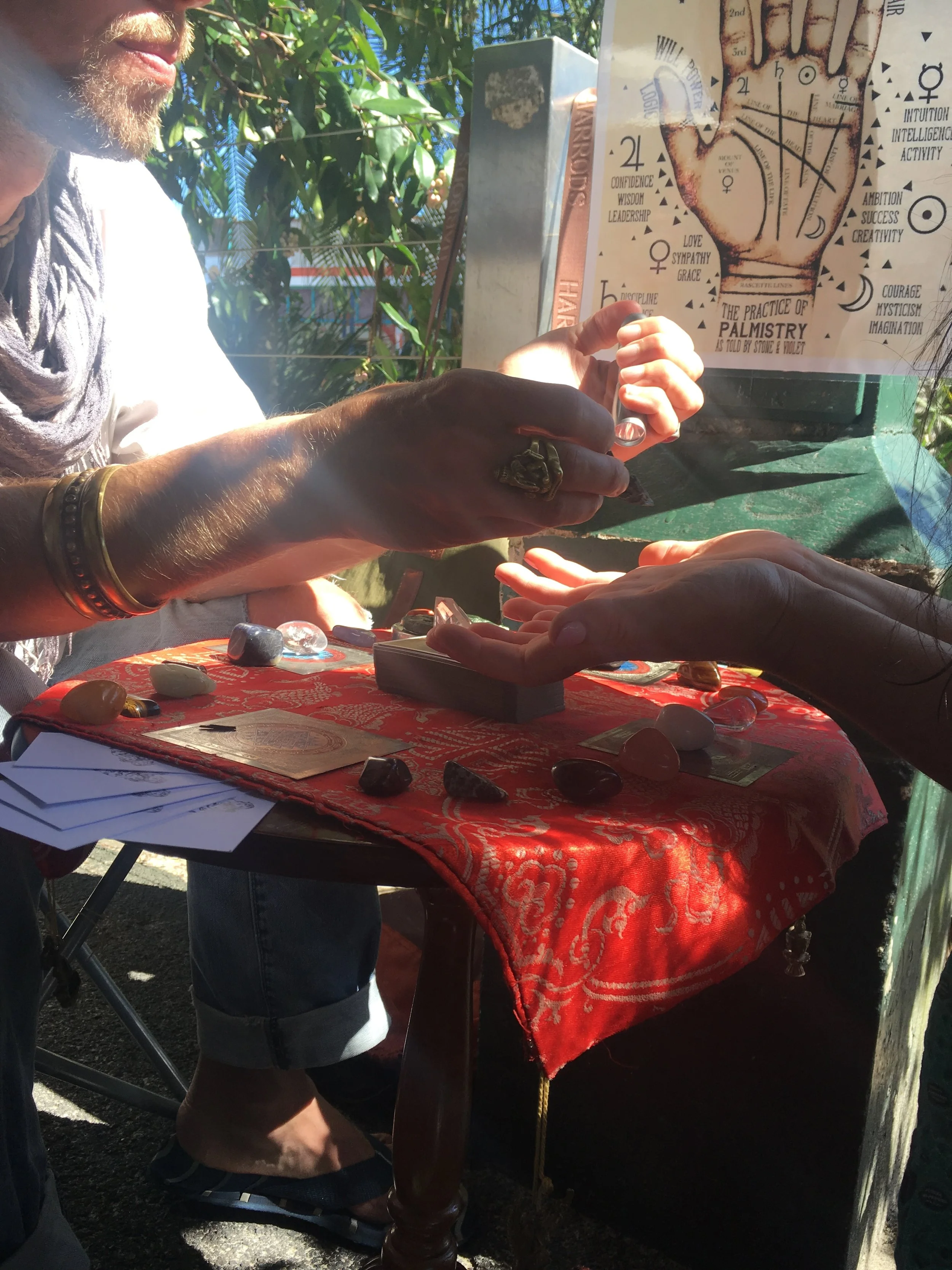 Palm reading setup with a person giving a reading. Red tablecloth with crystals. Palmistry chart displayed. Outdoor setting with foliage.