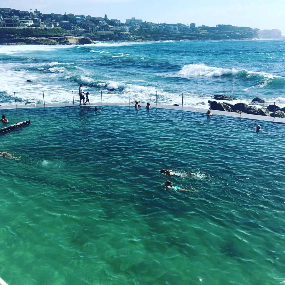 People swimming in an ocean pool by the seaside with waves in the background, surrounded by rocky coast.