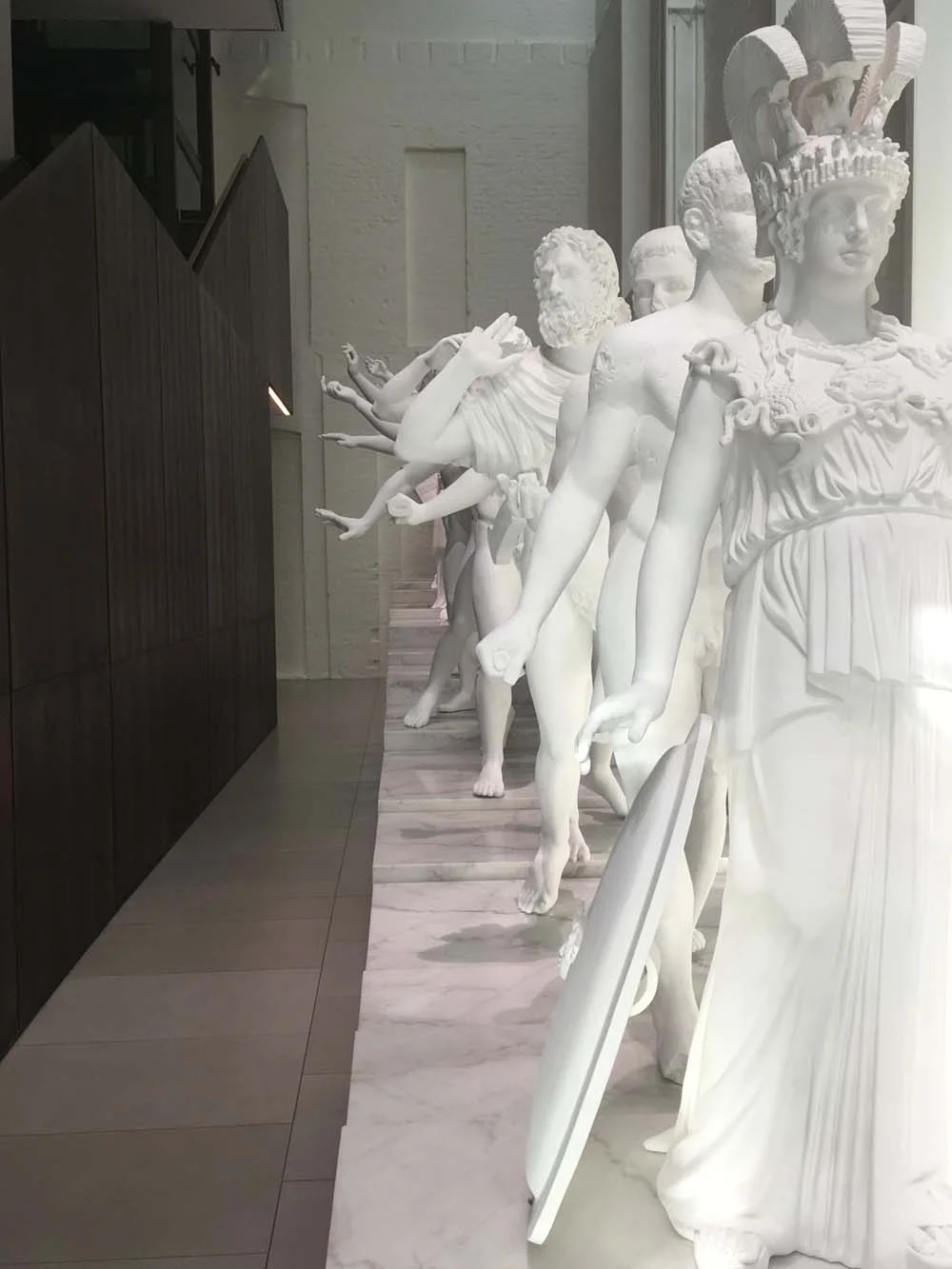 Row of classical white statues, indoors, on a marble surface, with modern architecture in the background.