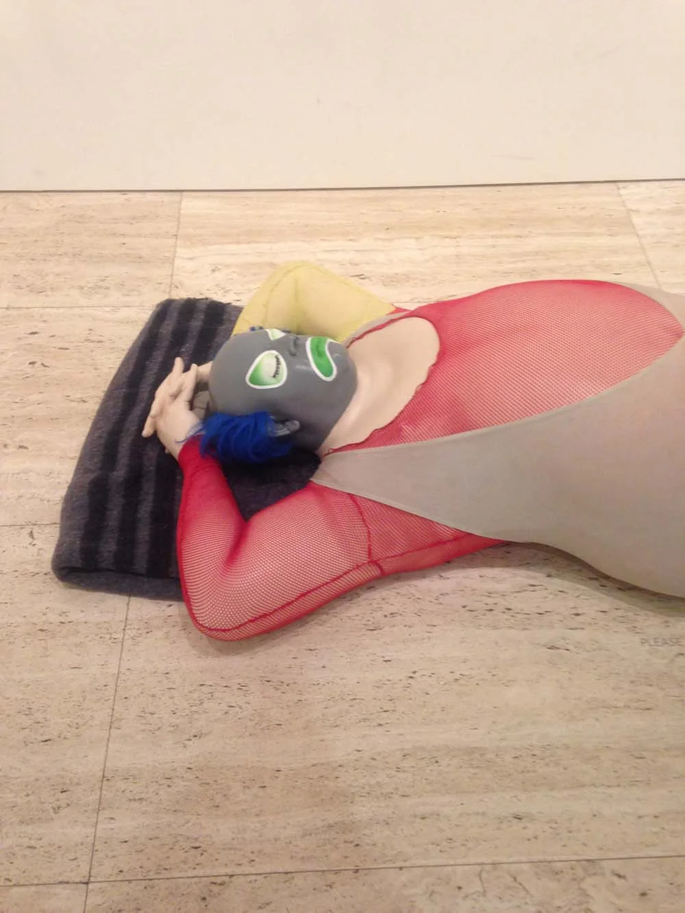 Artistic sculpture of a figure lying on a tiled floor, wearing a colorful bodysuit and a mask with green eye details, featuring a blue hairpiece.