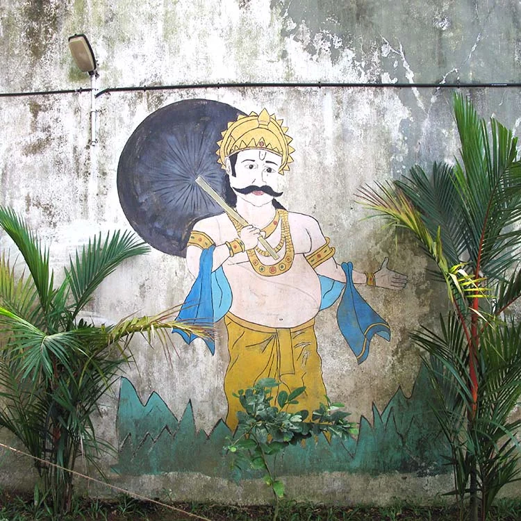Mural of a royal figure with a mustache, wearing traditional attire and holding an umbrella, painted on a wall with palm plants in the foreground.