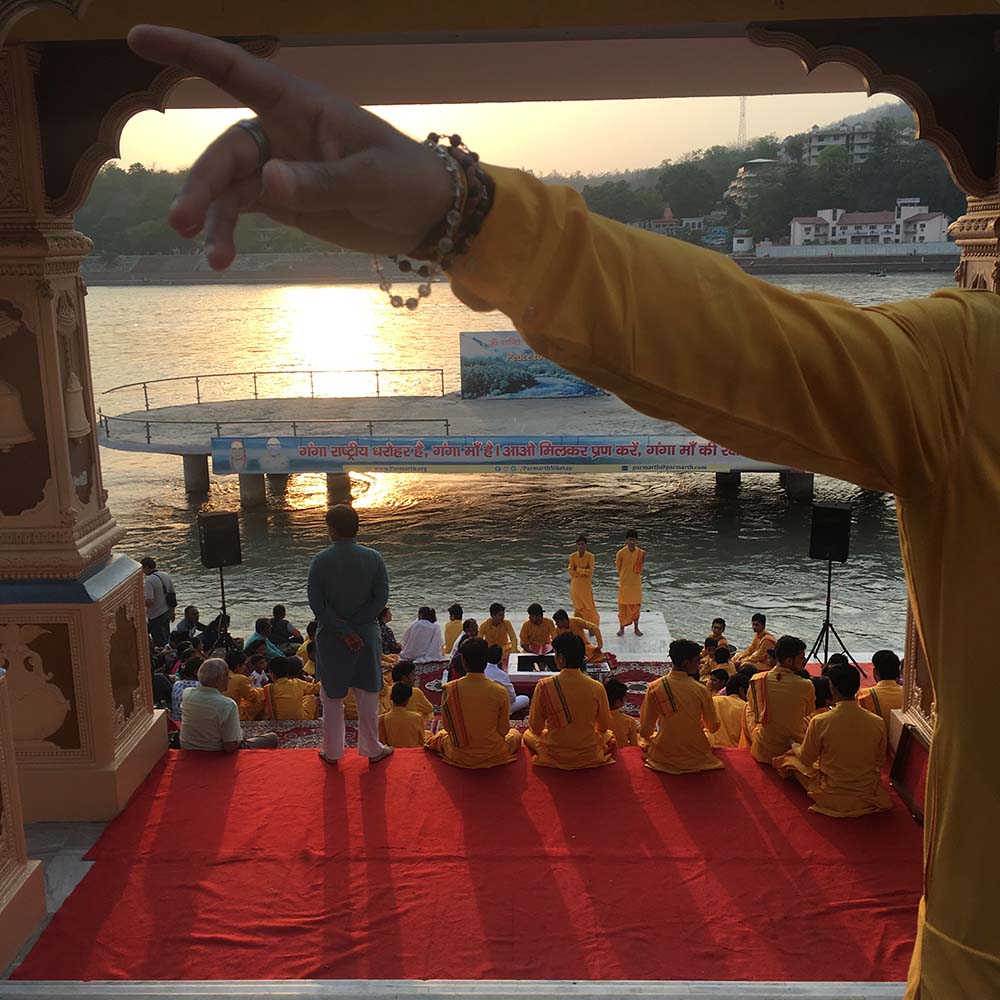 A group of people in traditional attire sitting on red carpet near a river during sunset.