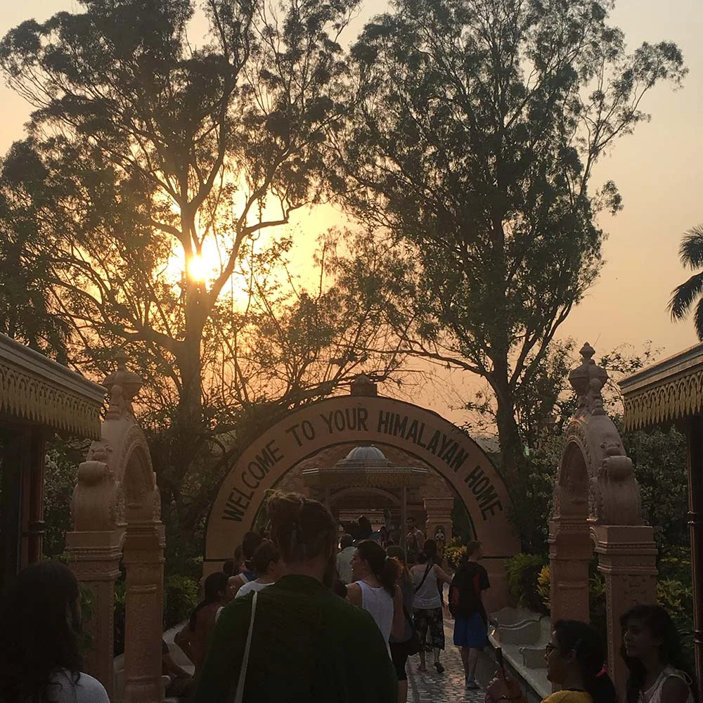 A group of people walking towards an archway with the text 'Welcome to Your Himalayan Home.' The sun is setting behind tall trees, creating a serene backdrop. The area is surrounded by decorative stone pillars.