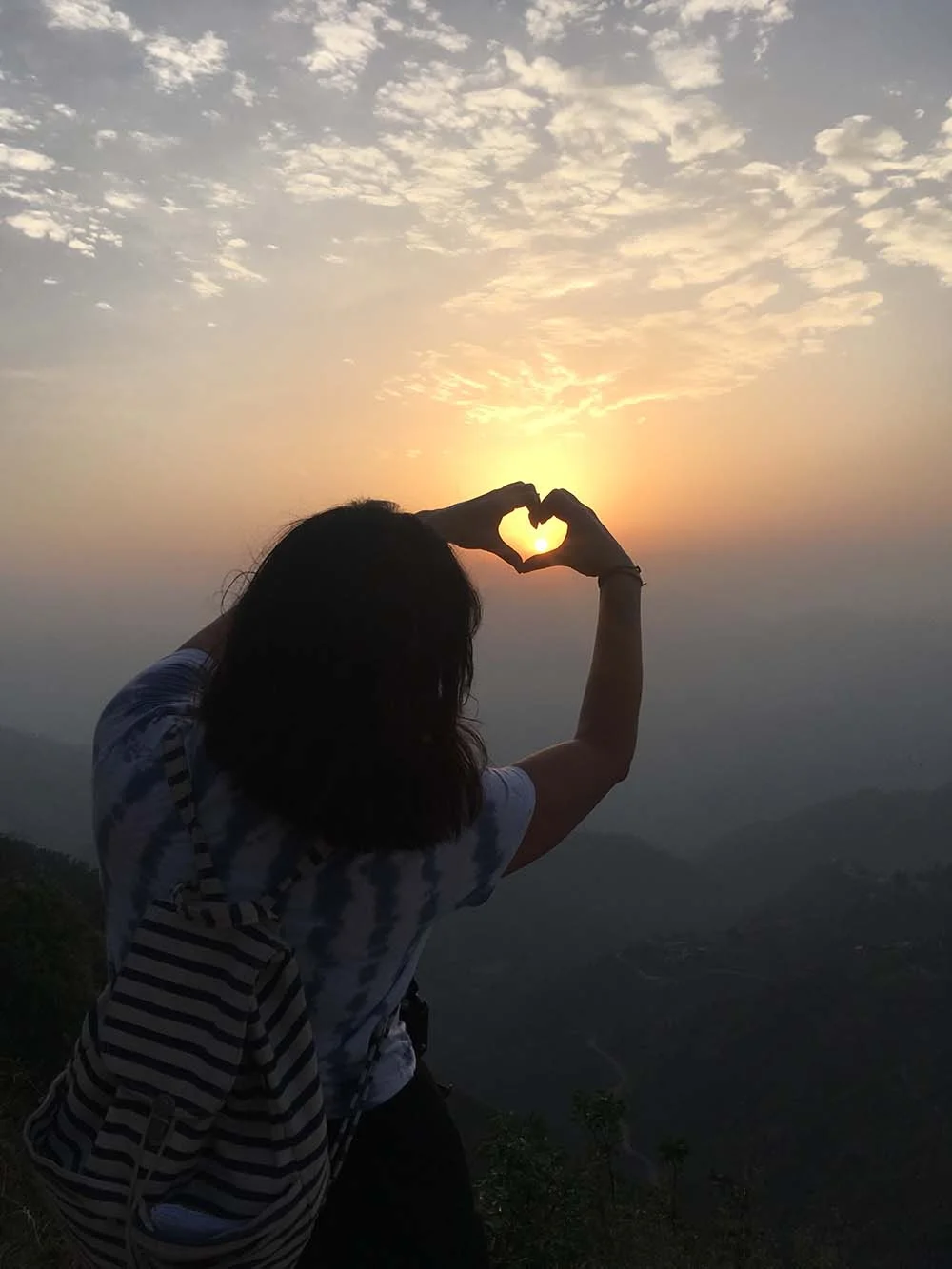 Person forming heart shape with hands against a sunrise or sunset with cloudy sky and mountains in the background.