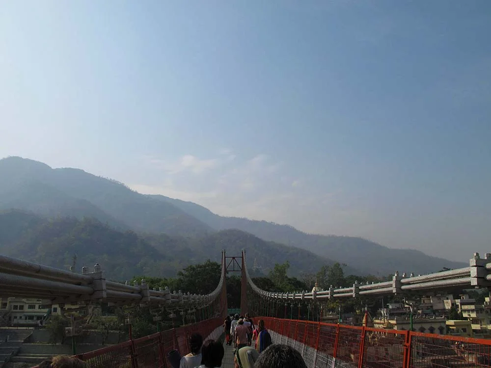 Suspension bridge with people walking across, mountains in the background, and hazy sky.