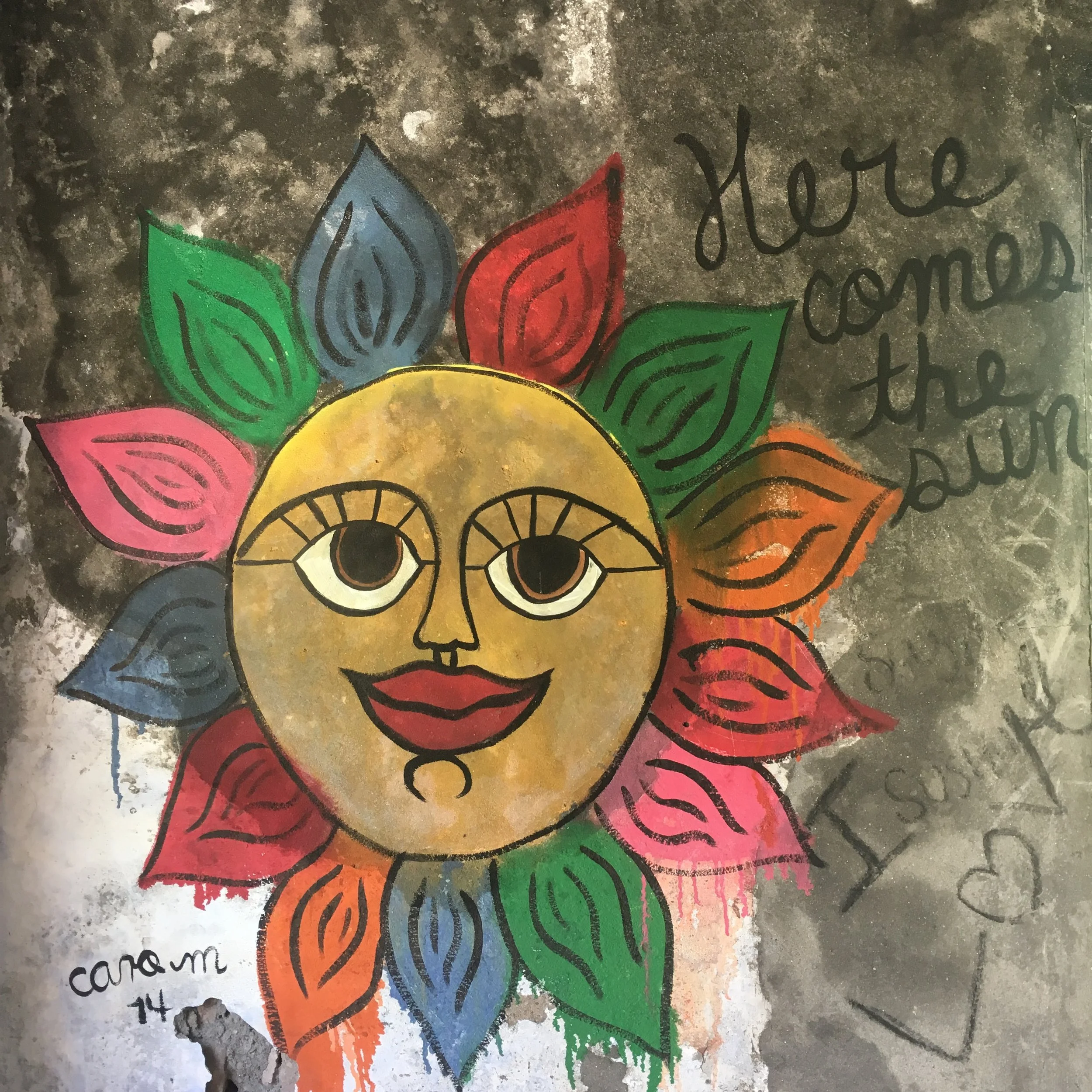 Colorful graffiti of a smiling sun with leaf-shaped rays in various colors on a textured wall, accompanied by the text 'Here comes the sun.'