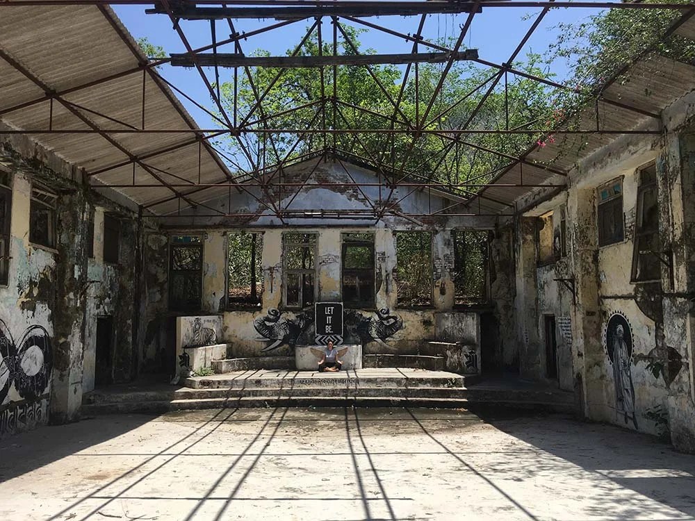 Abandoned building with graffiti and open roof structure, trees visible through open ceiling, sunlight creating shadows on concrete floor.