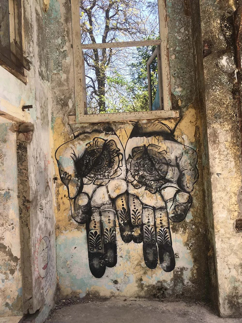 Artistic mural of detailed hands with henna designs on a weathered wall next to an open window, with a tree visible outside.