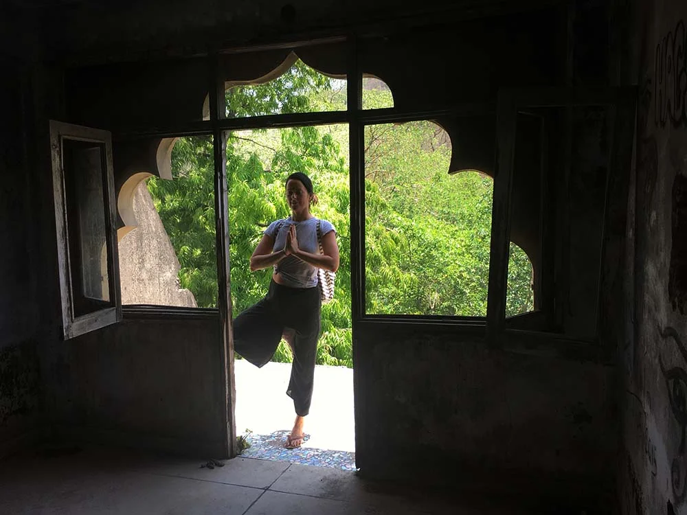 Person practicing yoga in front of an ornate window, framed by lush greenery outside.