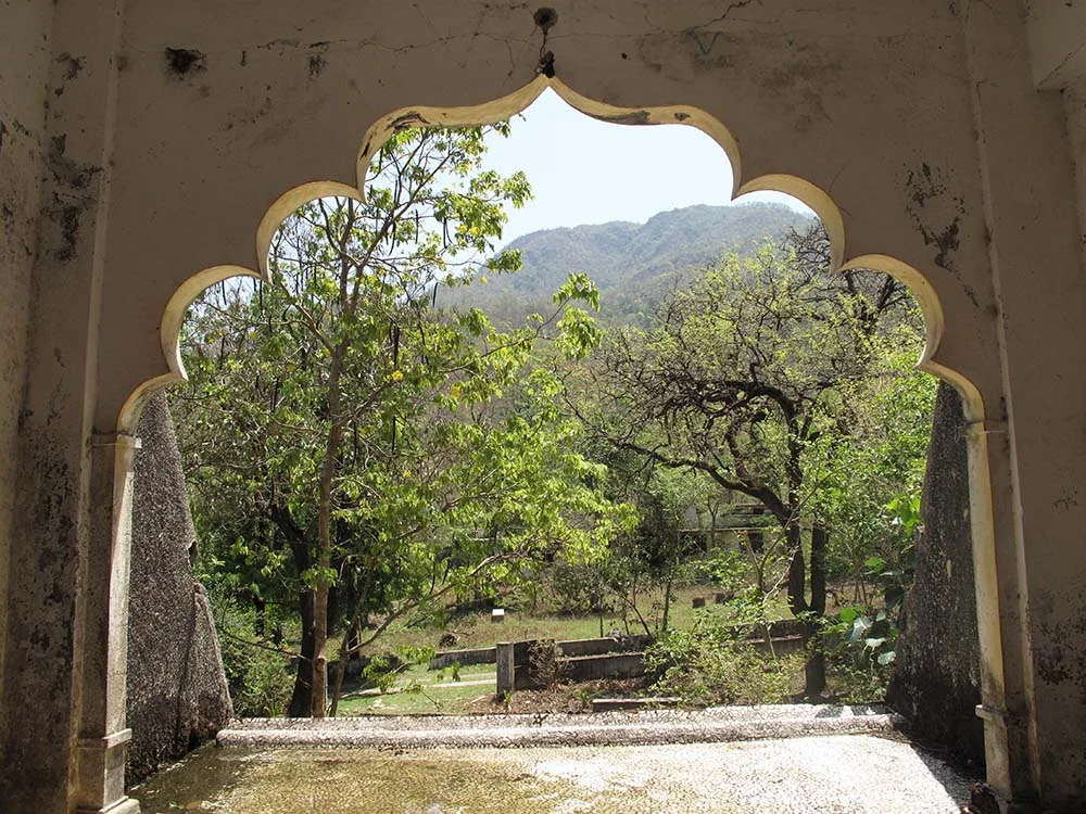 View through arched window of stone structure towards lush greenery and distant hills.