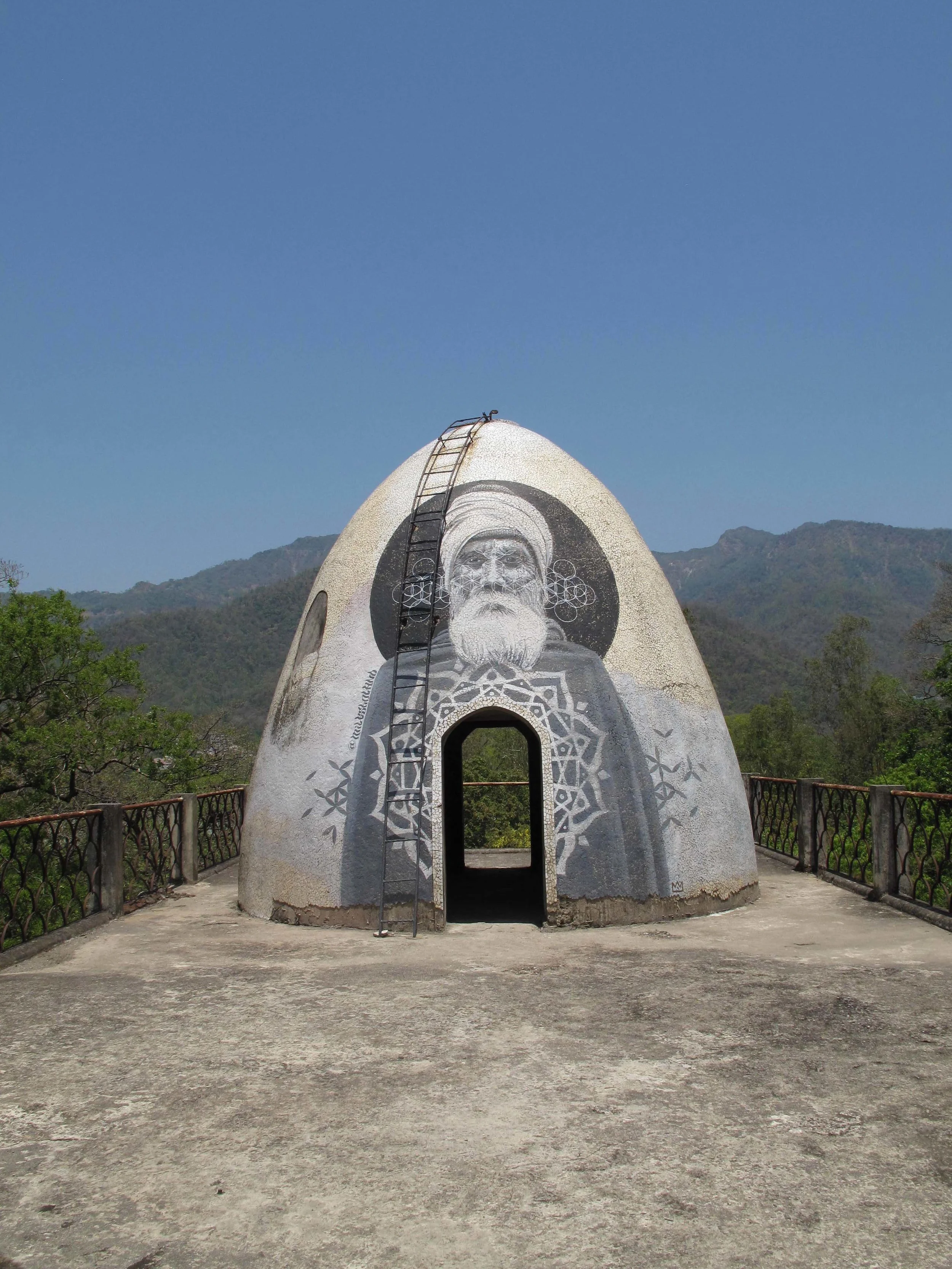 Dome-shaped structure with a mural of an elderly man, featuring a white beard and turban, with a ladder leaning against it, surrounded by a rail overlooking greenery and mountains under a clear blue sky.