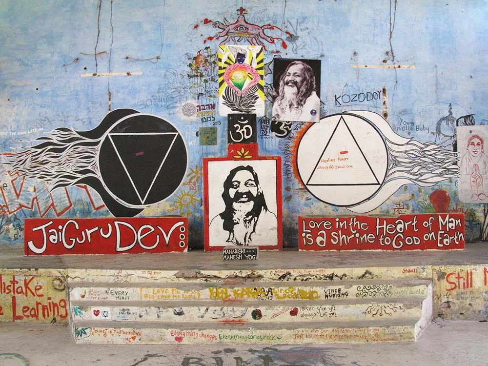 Colorful wall mural with spiritual symbols, text 'Jai Guru Dev', portraits, and quotes, including 'Love in the heart of man is a shrine to God on Earth'.