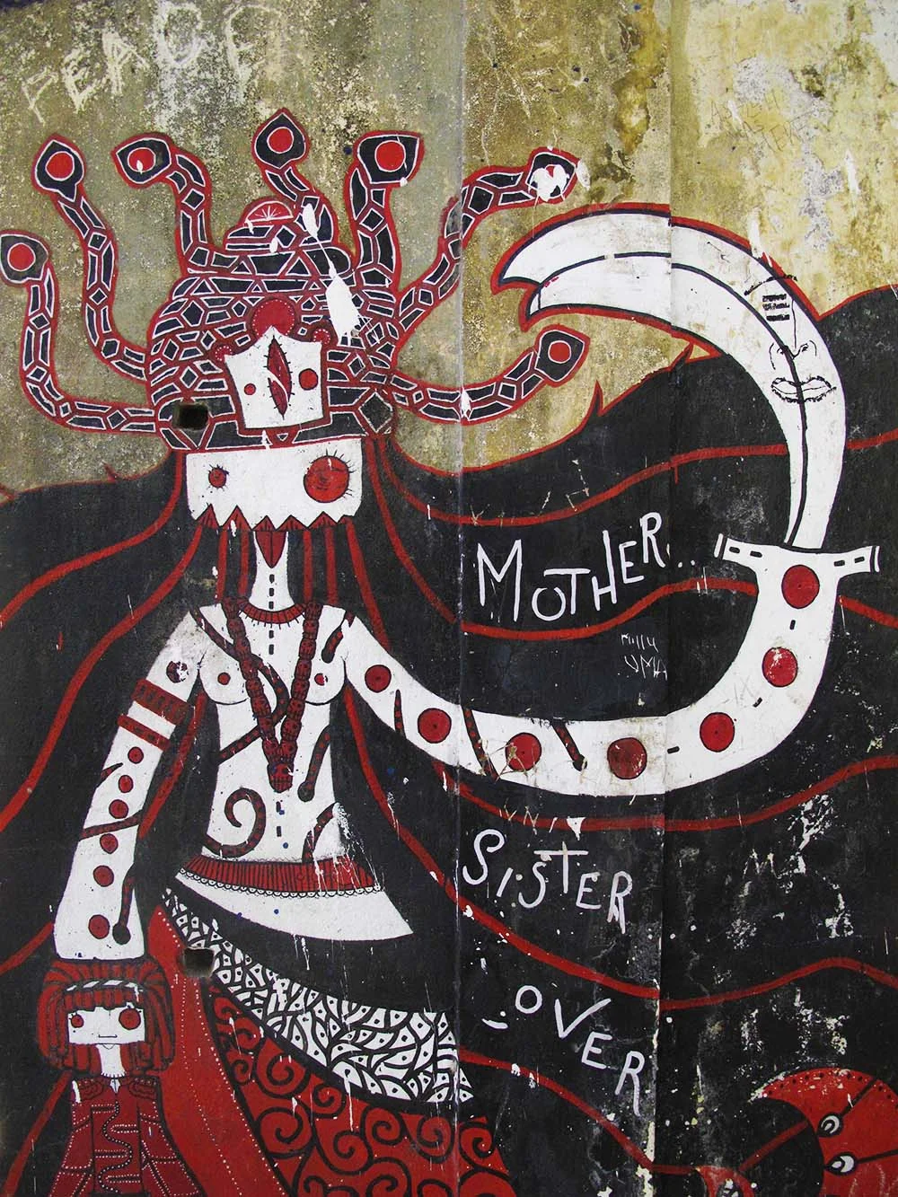 Colorful street art mural depicting a stylized humanoid figure with a headdress resembling snakes, holding a sickle in one hand. The artwork features red, black, and white colors with words "Mother," "Sister," and "Lover" painted across it, along wit