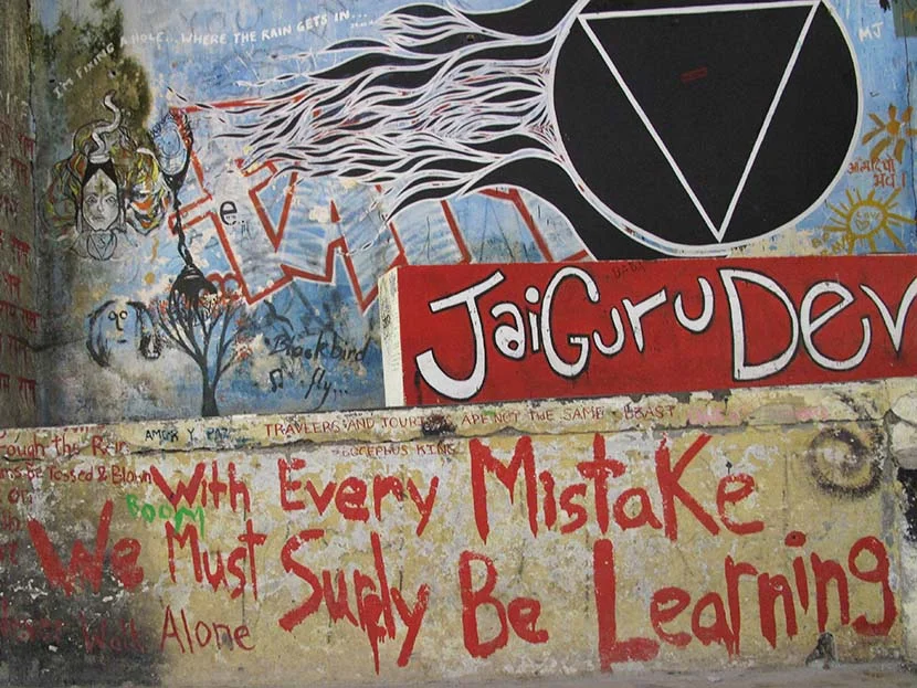 Wall with colorful graffiti art, including text "Jai Guru Dev" and "With Every Mistake We Must Surely Be Learning," along with various abstract designs and drawings.
