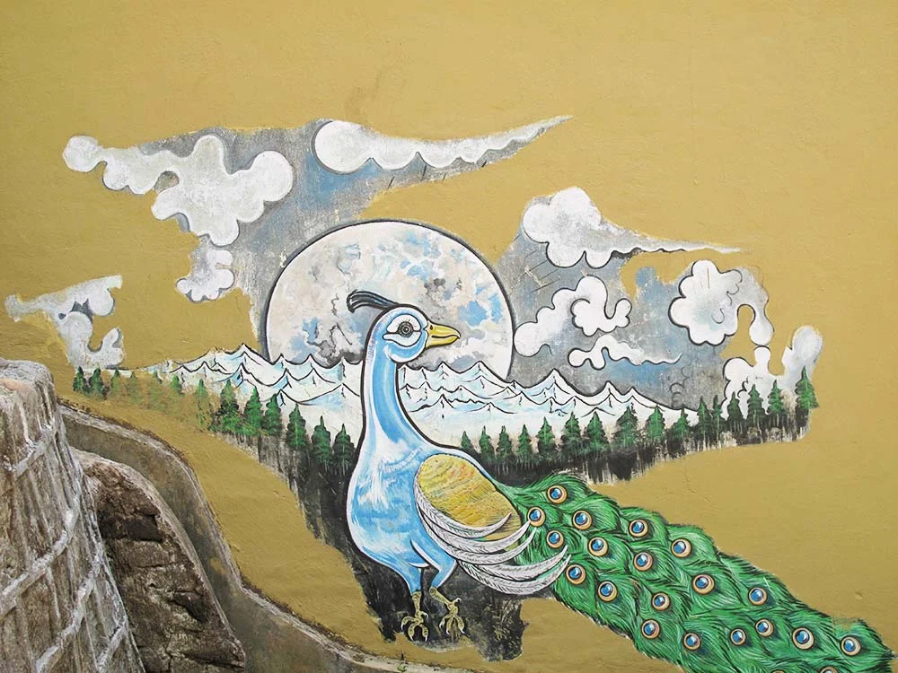 Mural of a stylized peacock with a colorful tail in front of snow-capped mountains and clouds on a yellow wall.