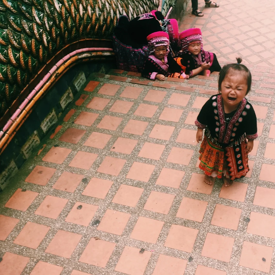 Young child wearing traditional clothing standing on tiled steps, with two other children and an adult in similar attire seated on the stairs in the background.