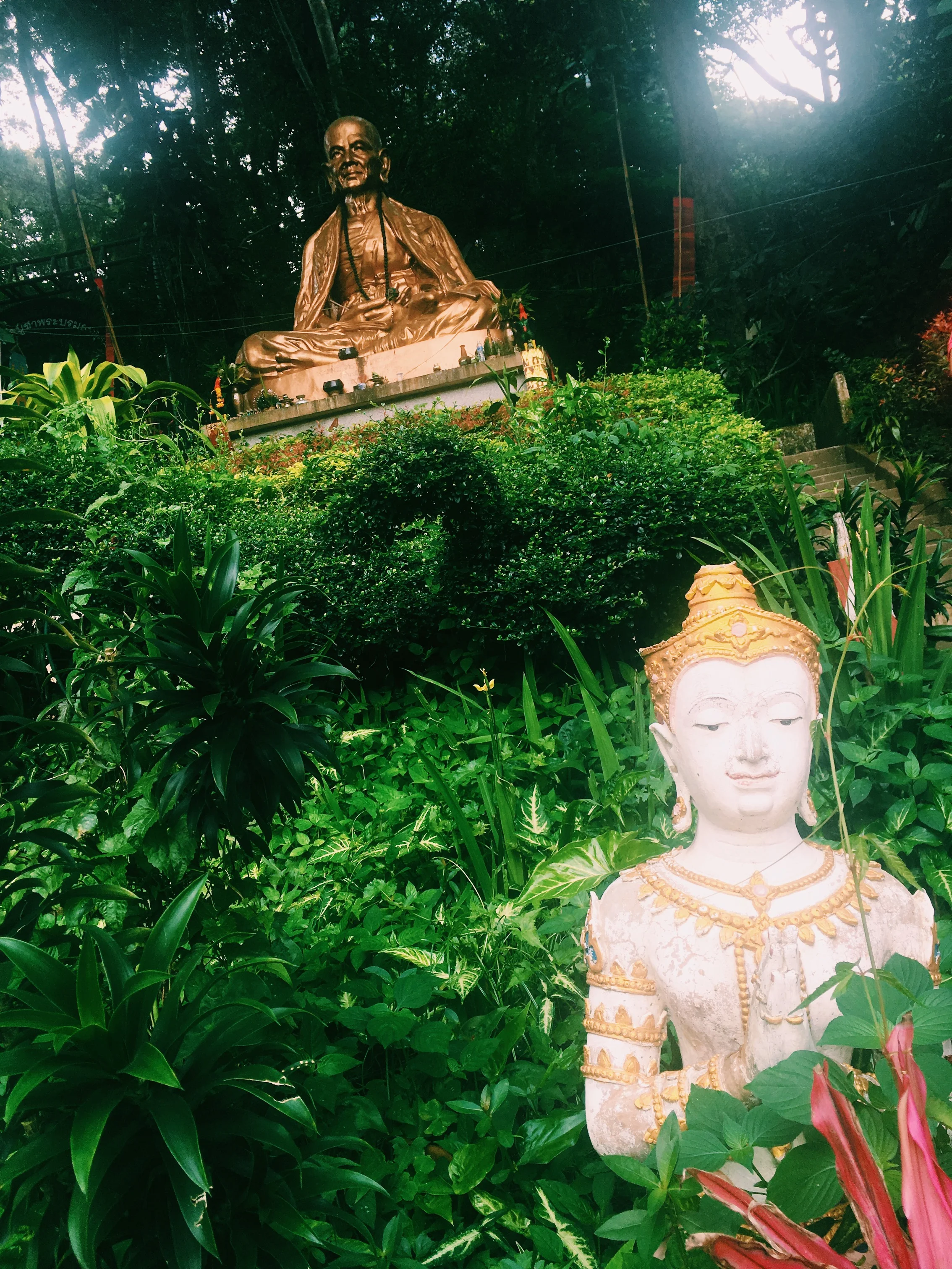 Outdoor setting with a large bronze statue of a seated person and a smaller stone statue in a prayer position surrounded by lush greenery.