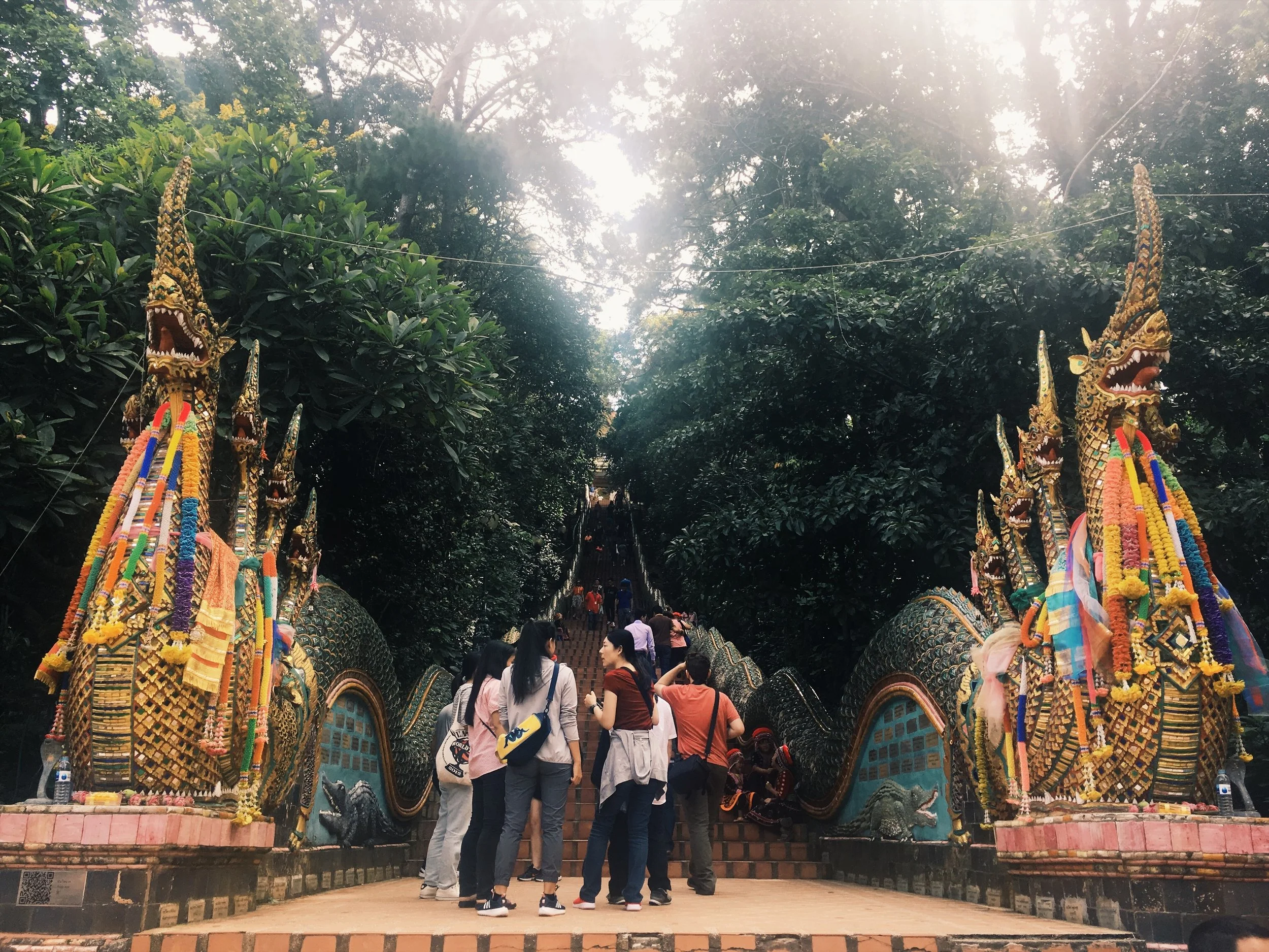 Group of people ascending a staircase adorned with ornate Naga serpent statues at a temple surrounded by lush greenery.